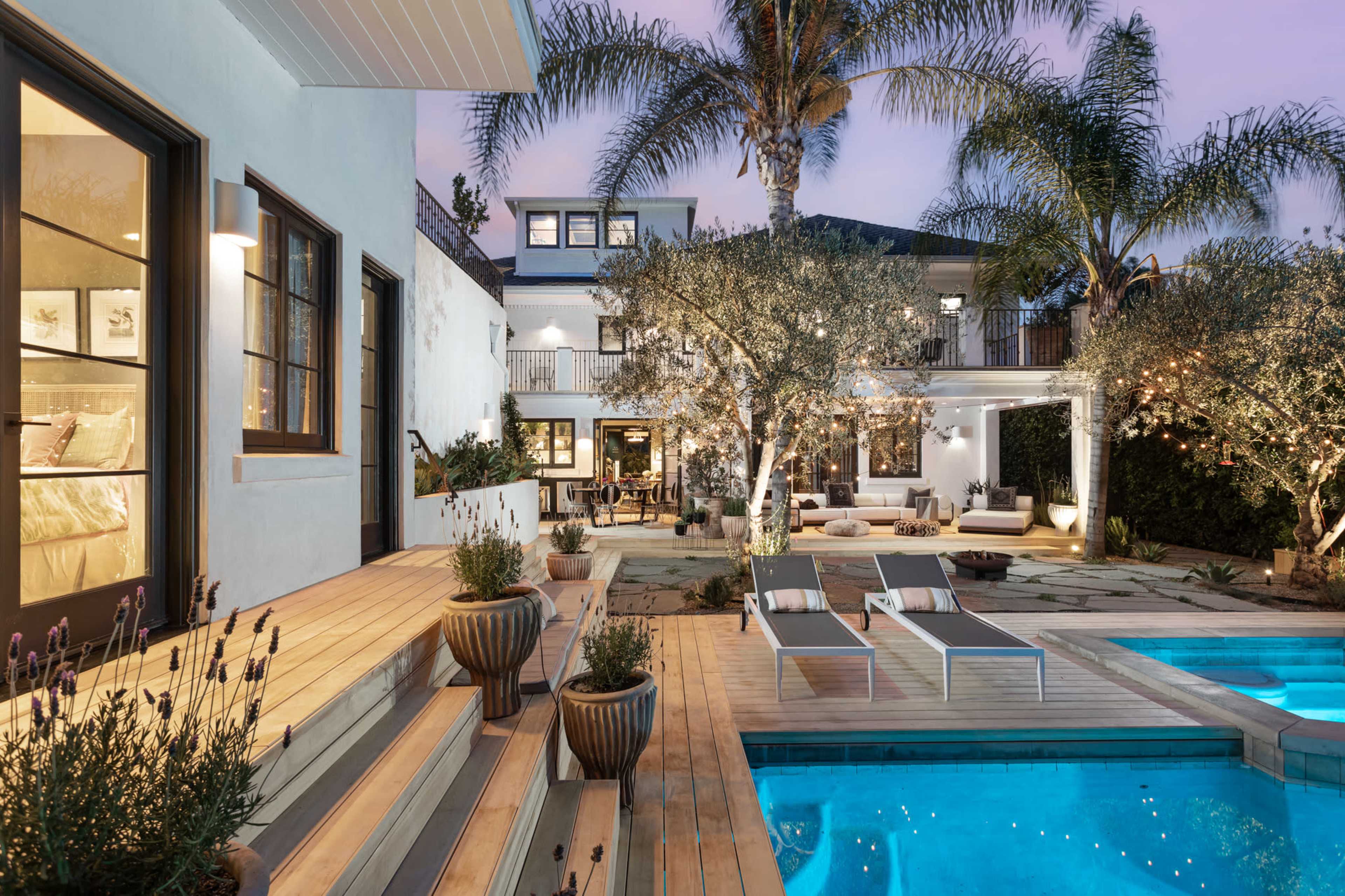 The image shows a well-designed backyard with a pool, lounge chairs, and surrounding greenery, illuminated by evening lights.