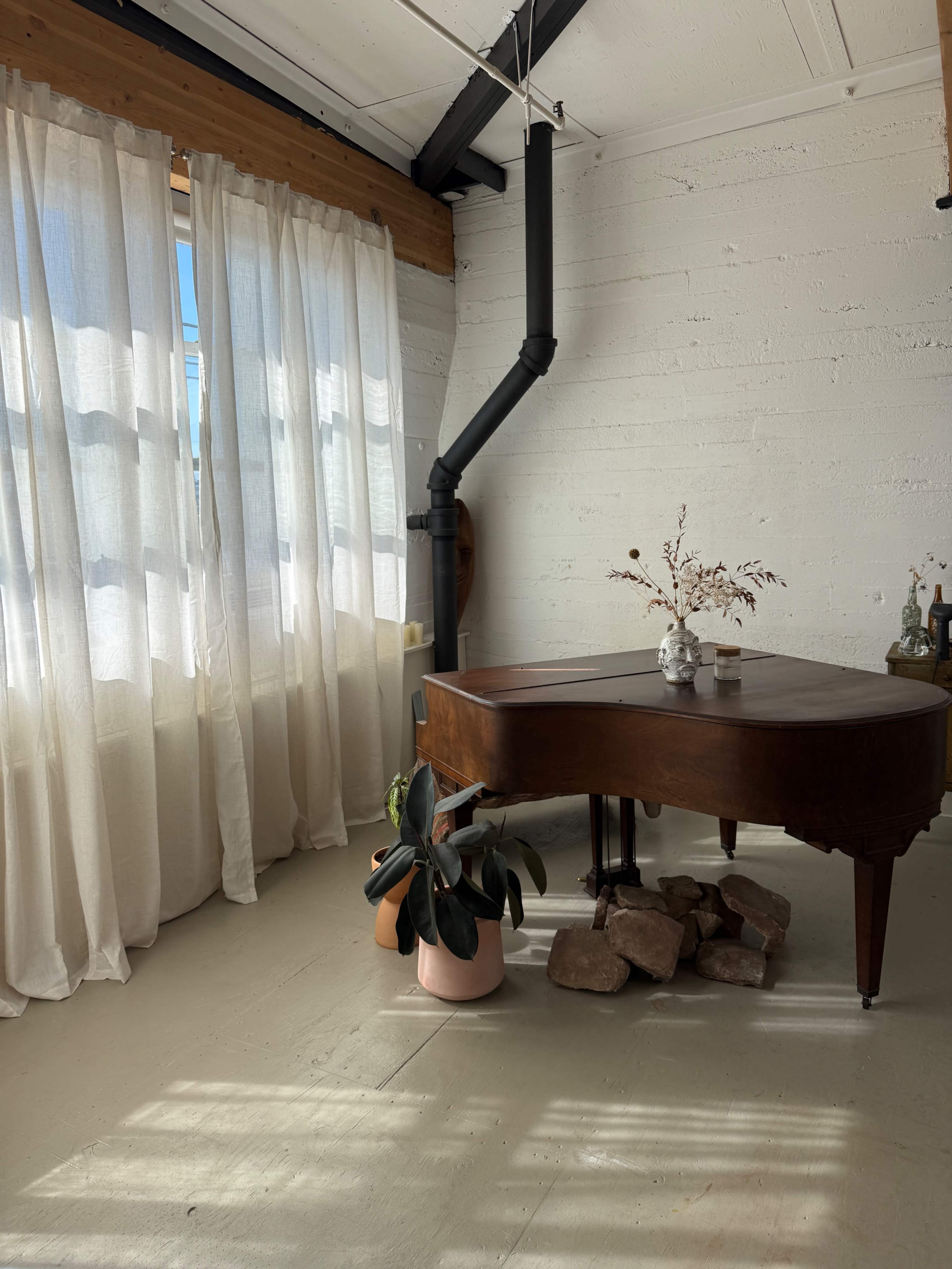 A grand piano sits on the floor near a window, with sheer curtains filtering light and a potted plant beside it on a stack of rocks.