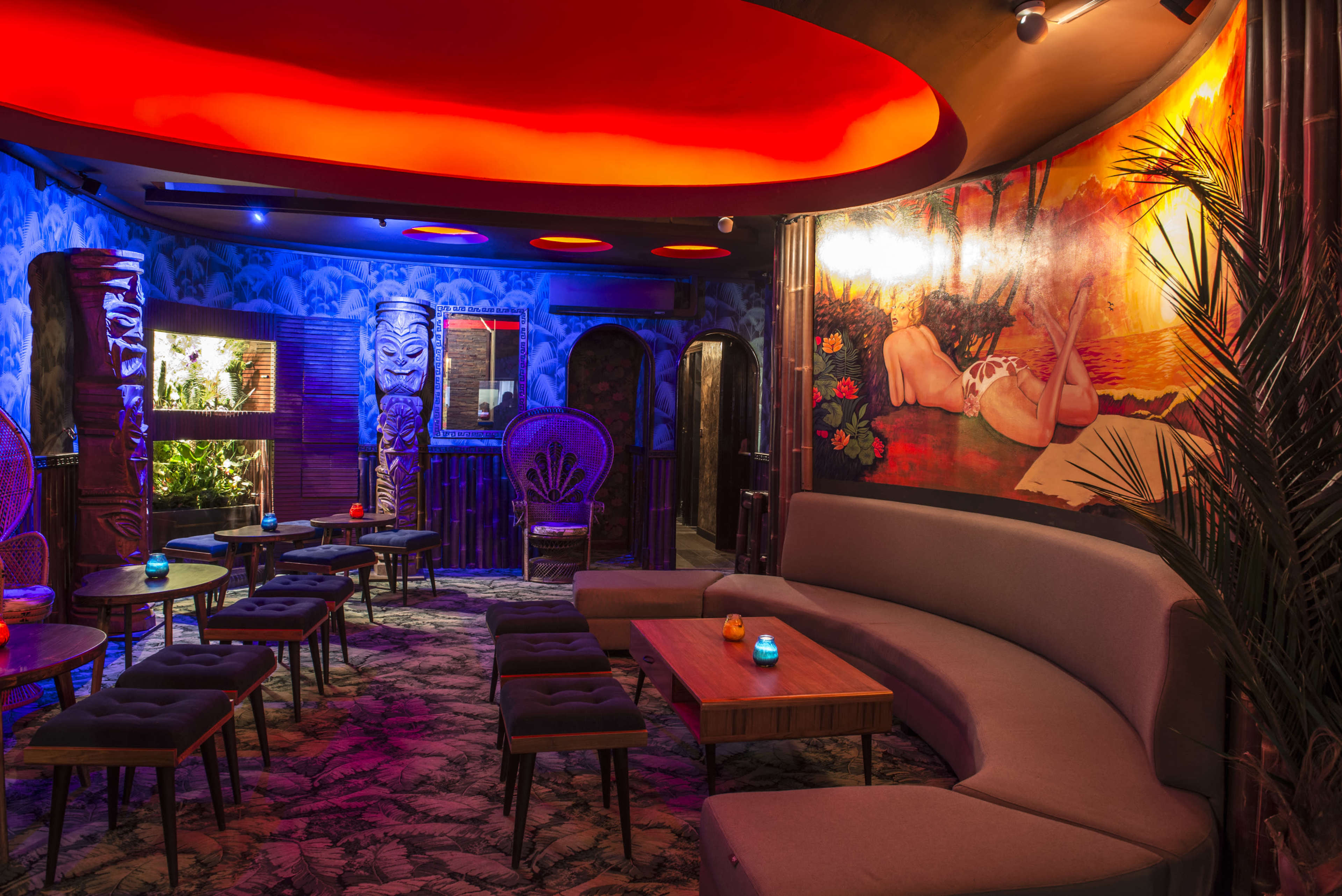 The image shows a dimly lit lounge area with a curved sofa, wooden tables, and tropical-themed decor featuring vibrant artwork and tiki-style sculptures.