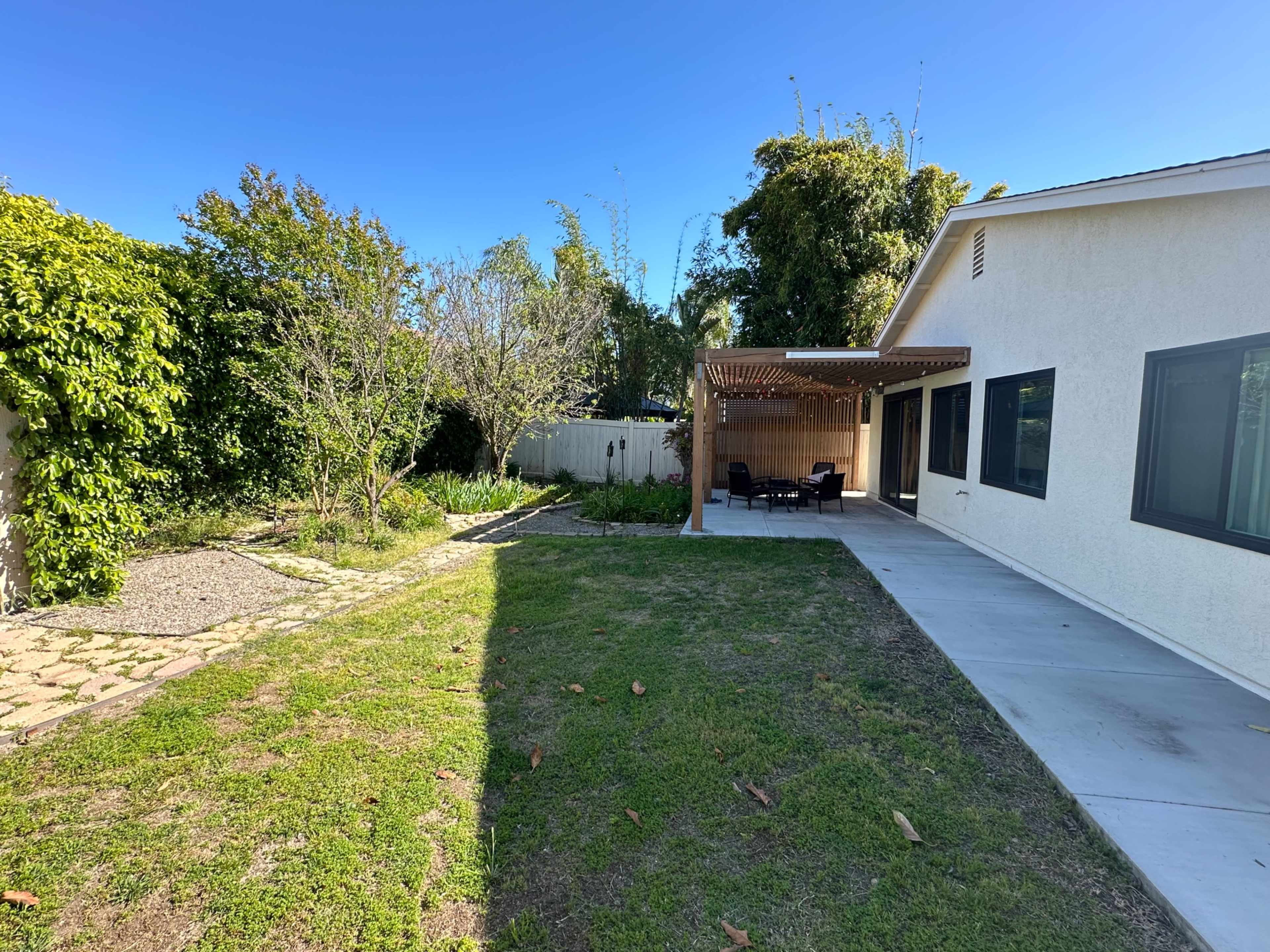A residential backyard with a paved area, grassy sections, a garden path, and a covered patio with chairs.