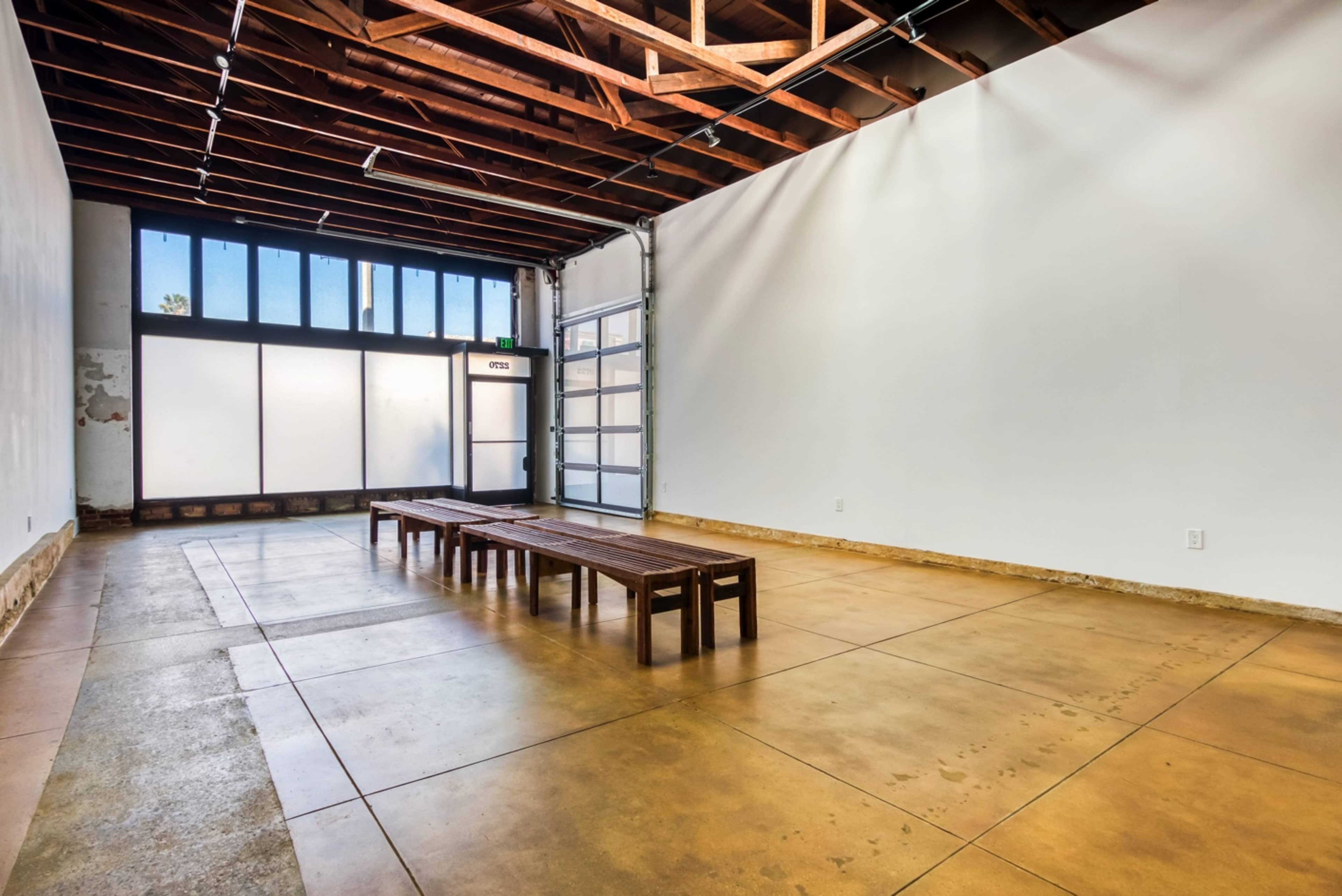 The image shows a spacious, empty room with a polished concrete floor and wooden beams overhead, featuring a long wooden table in the center and large windows on one side.
