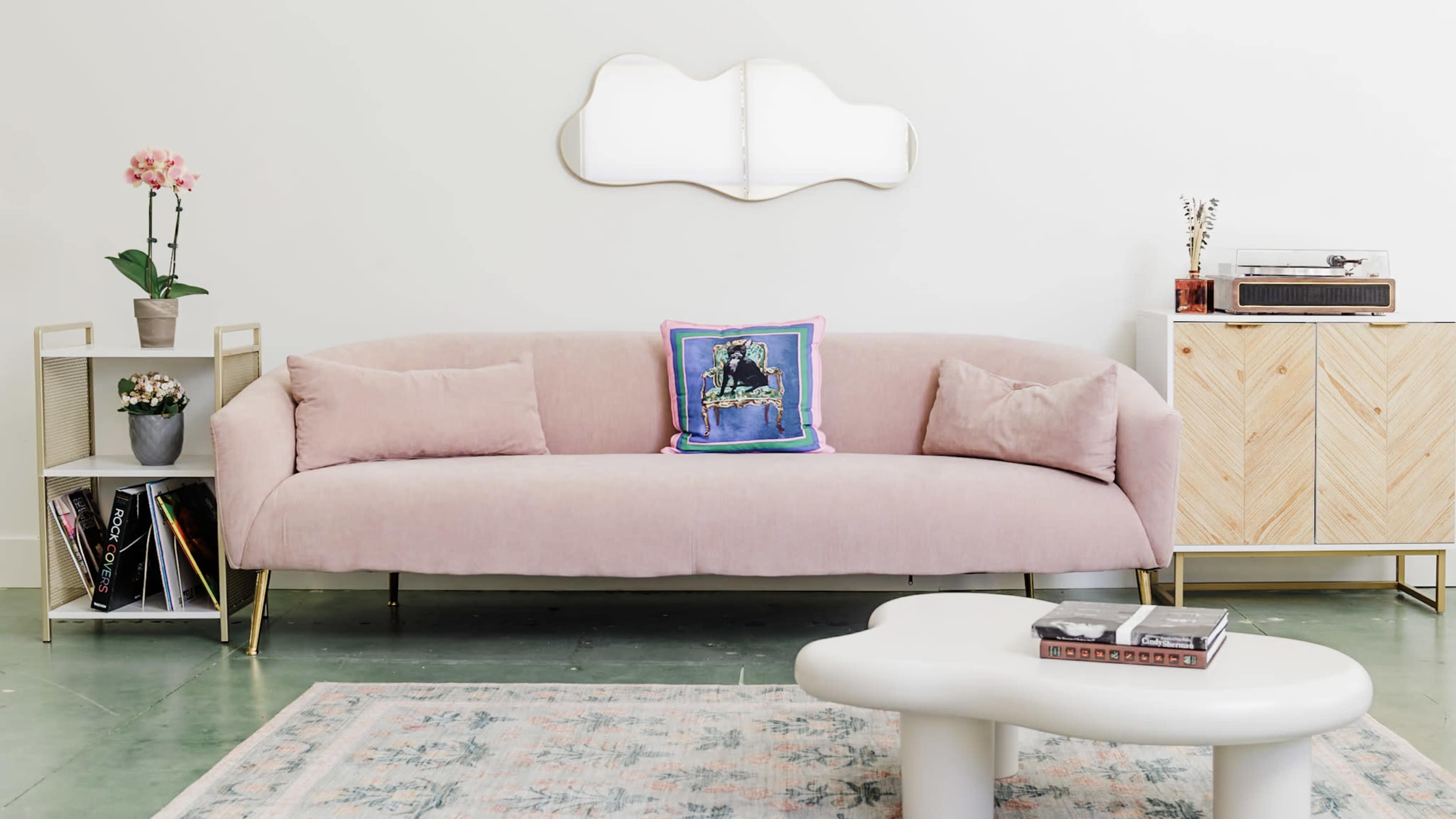 A light pink sofa with a decorative pillow, positioned in a room that features a coffee table, a side table with a plant, and a cabinet.