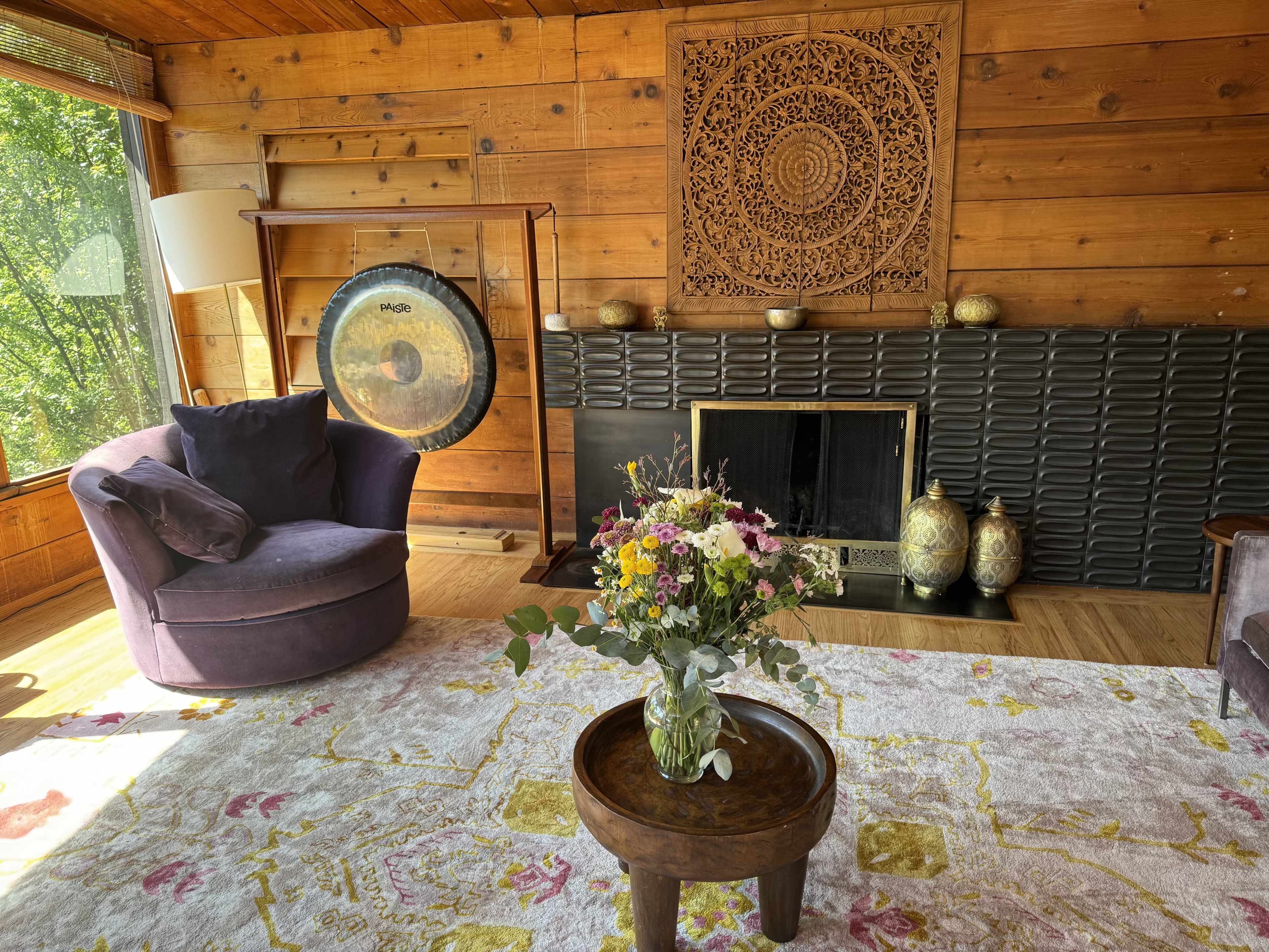 A cozy living room features a gong, a decorative wall panel, a floral arrangement on a wooden table, and wooden walls with large windows allowing natural light.