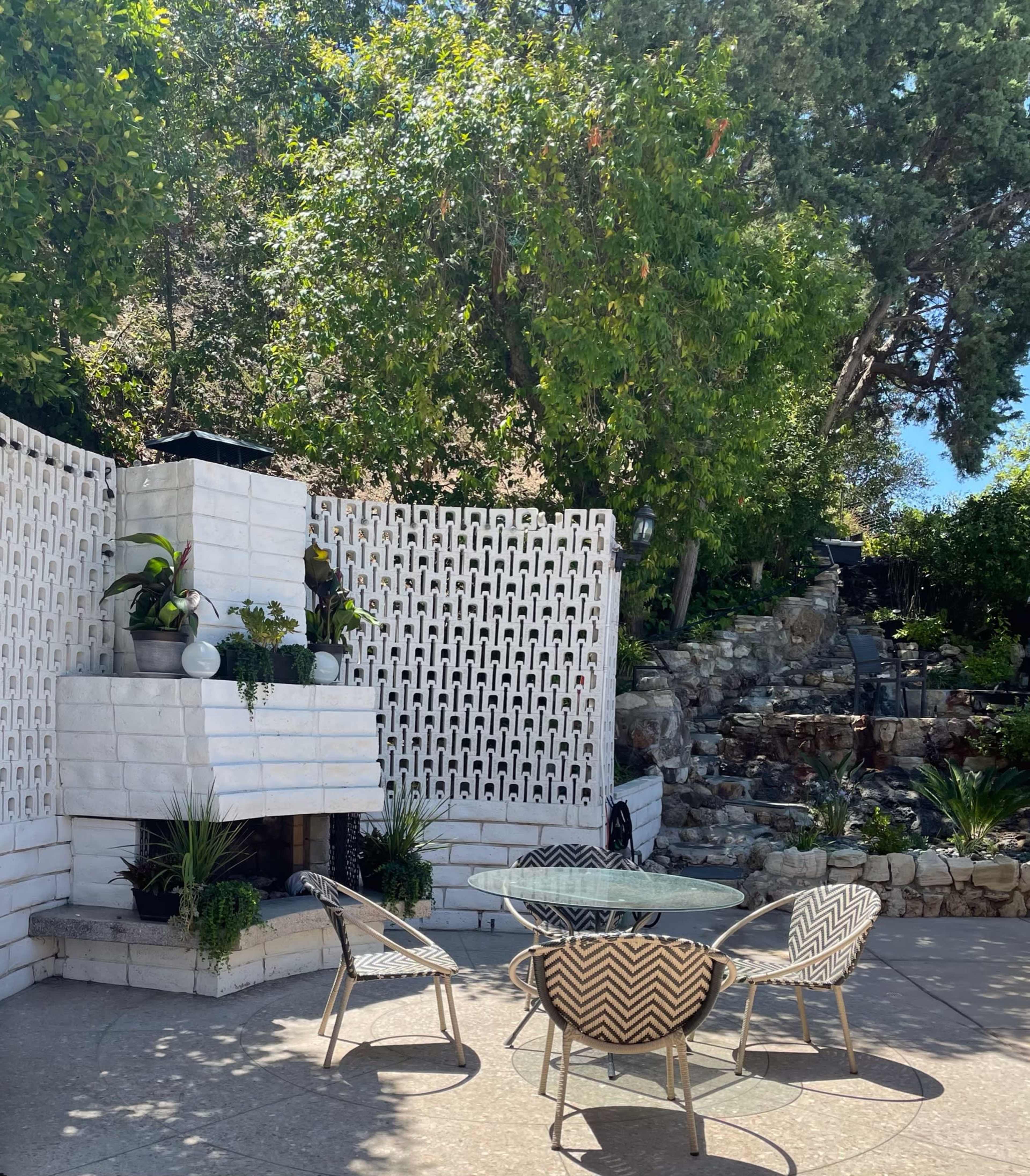 A patio features a stone fireplace and several chairs surrounded by greenery and a patterned wall.