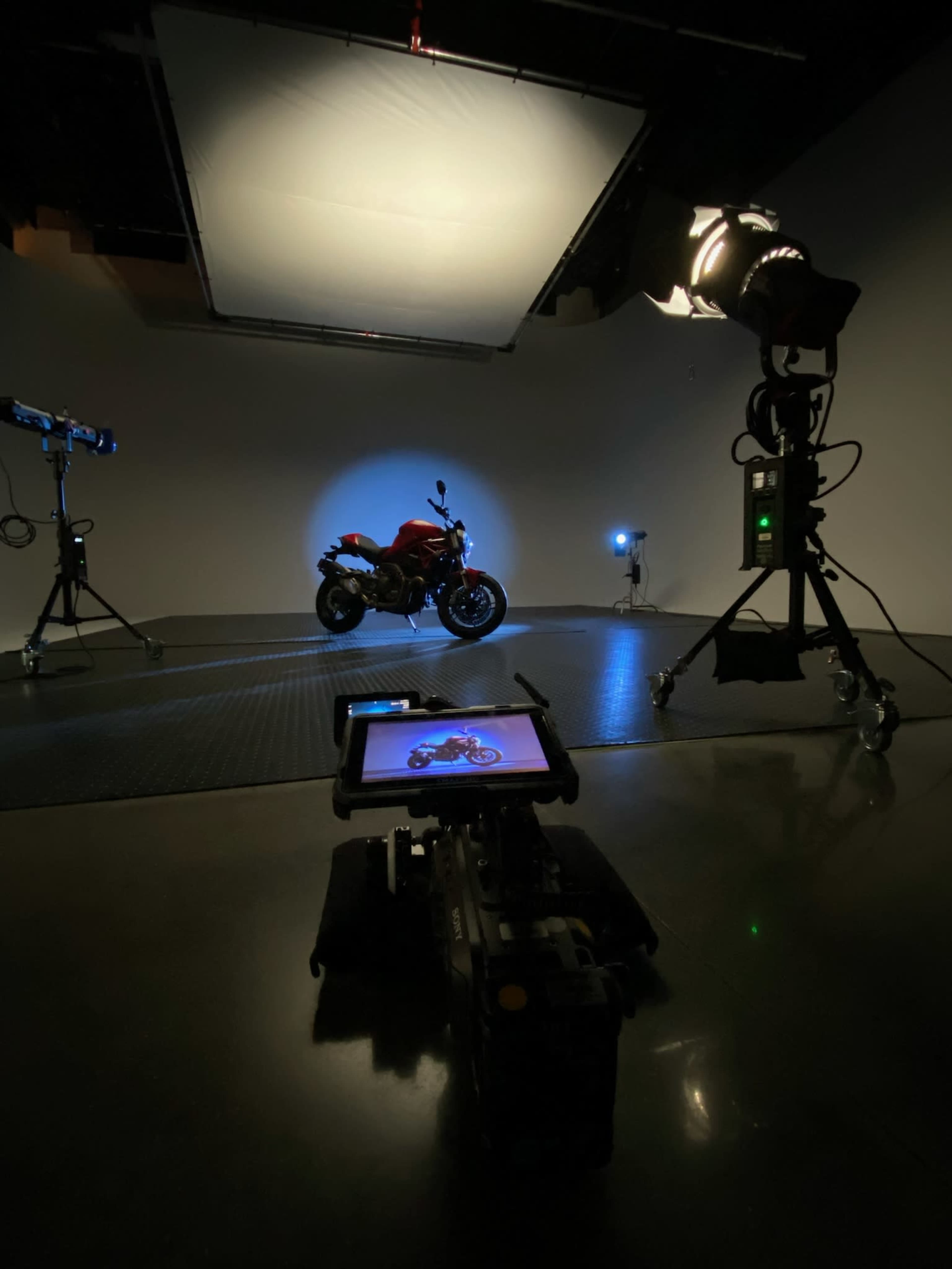 A red motorcycle is illuminated in a photo studio, surrounded by lighting equipment and a camera display showing an image of the bike.