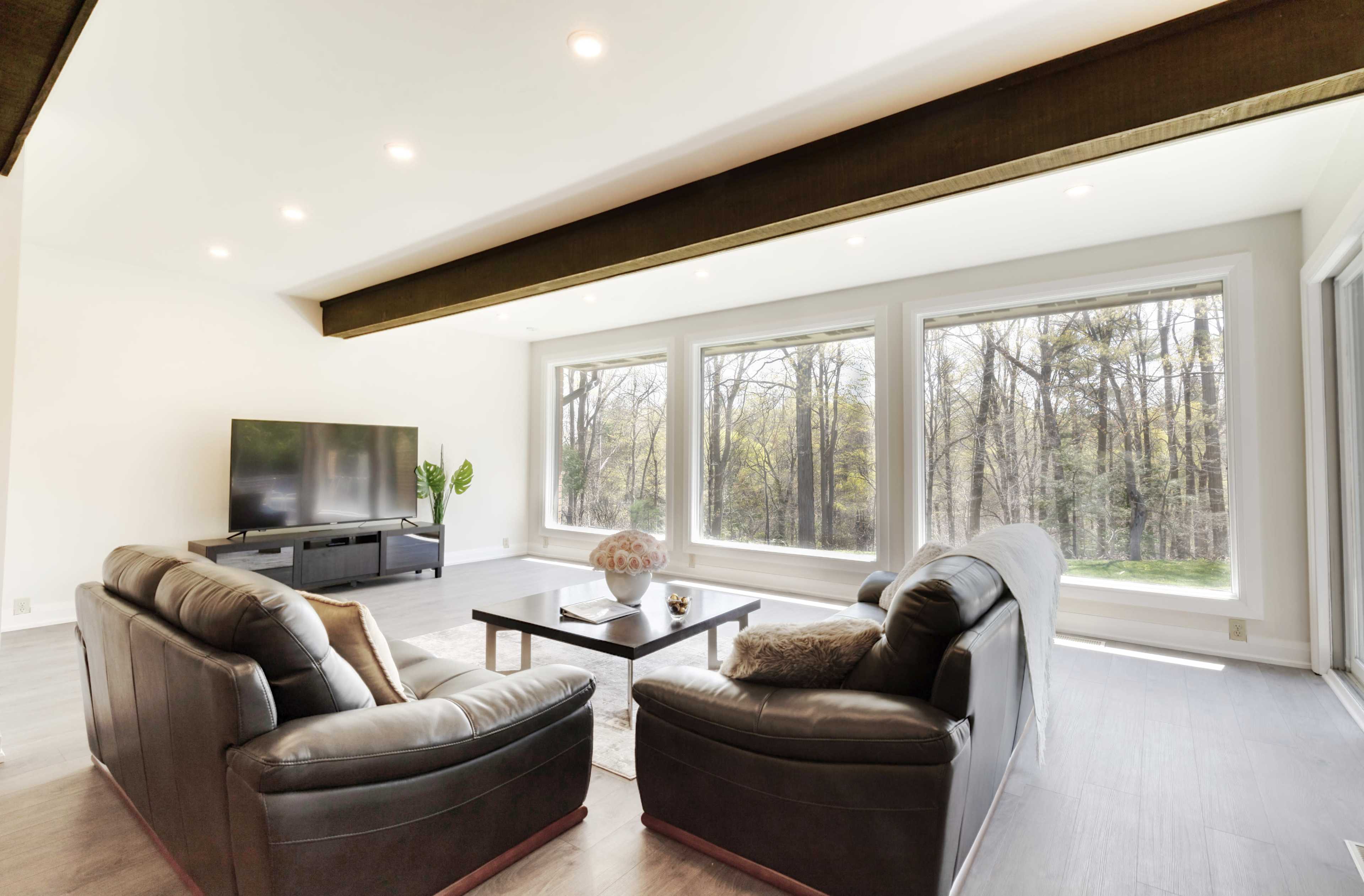 A modern living room features two leather sofas facing a coffee table, with a television and large windows showcasing a view of trees outside.