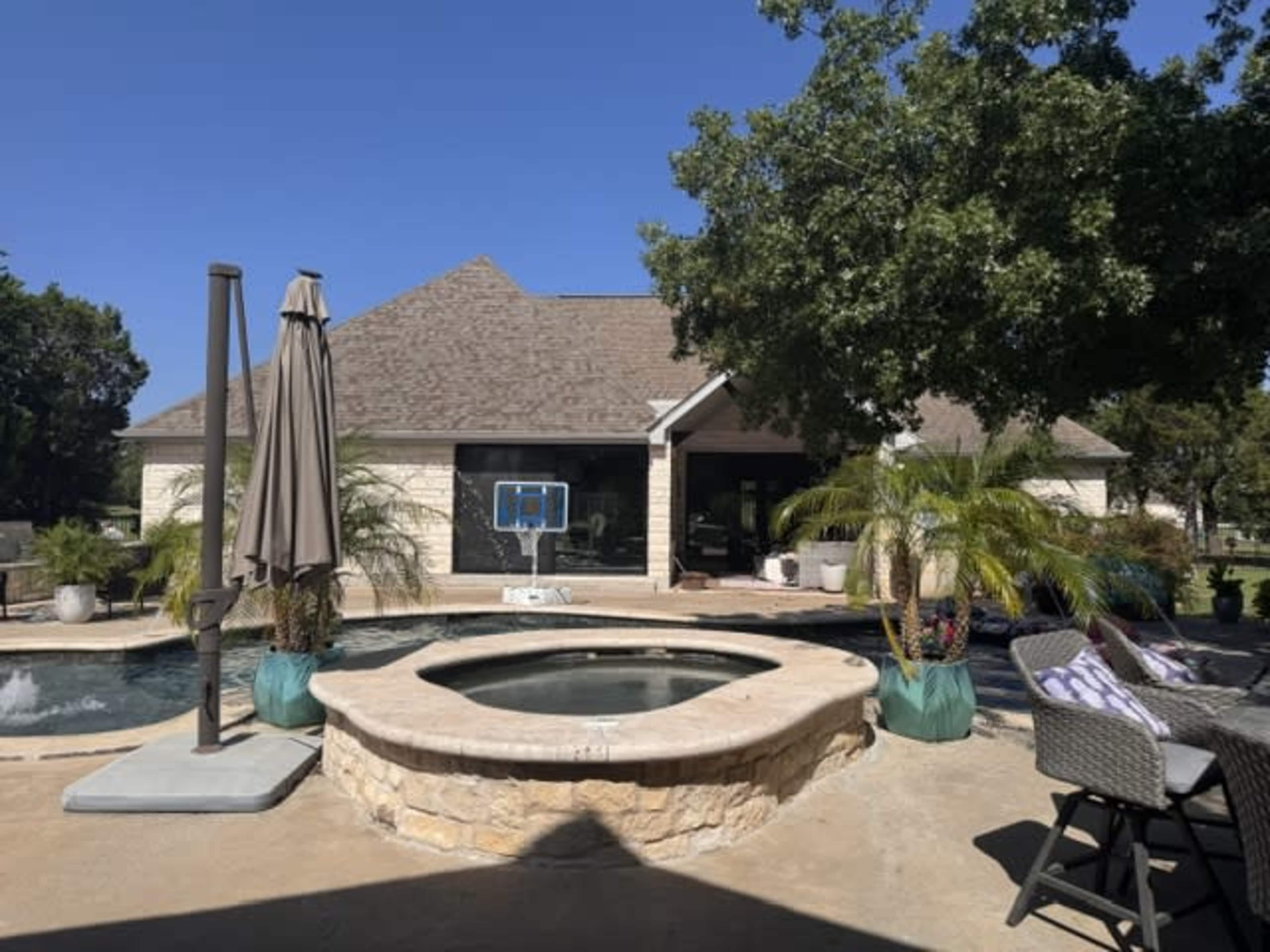 The image shows a backyard with a hot tub, a basketball hoop, and a patio area surrounded by trees and a house in the background.