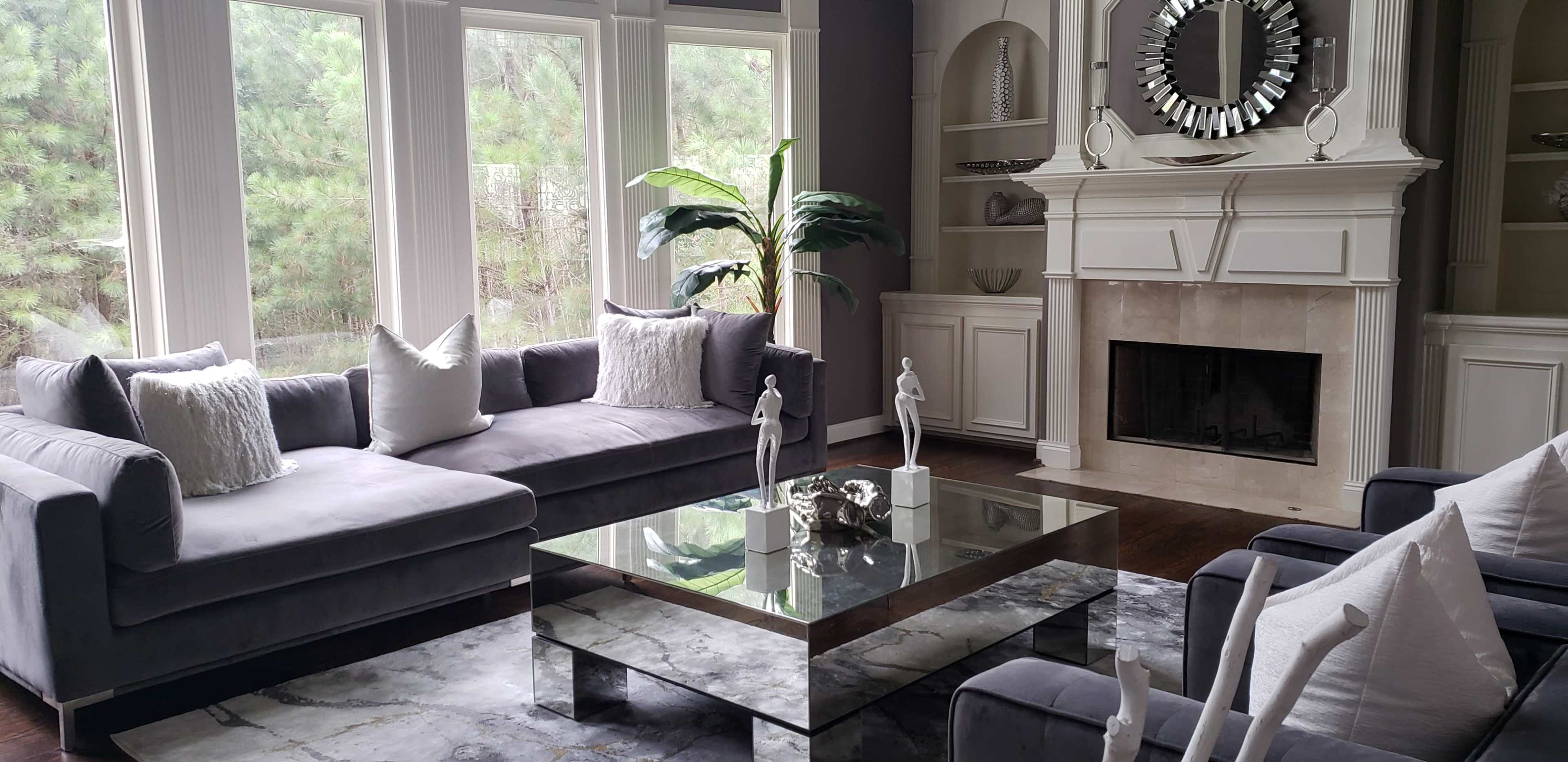 The image depicts a modern living room featuring a gray sectional sofa, a marble coffee table, decorative sculptures, and large windows with views of greenery.