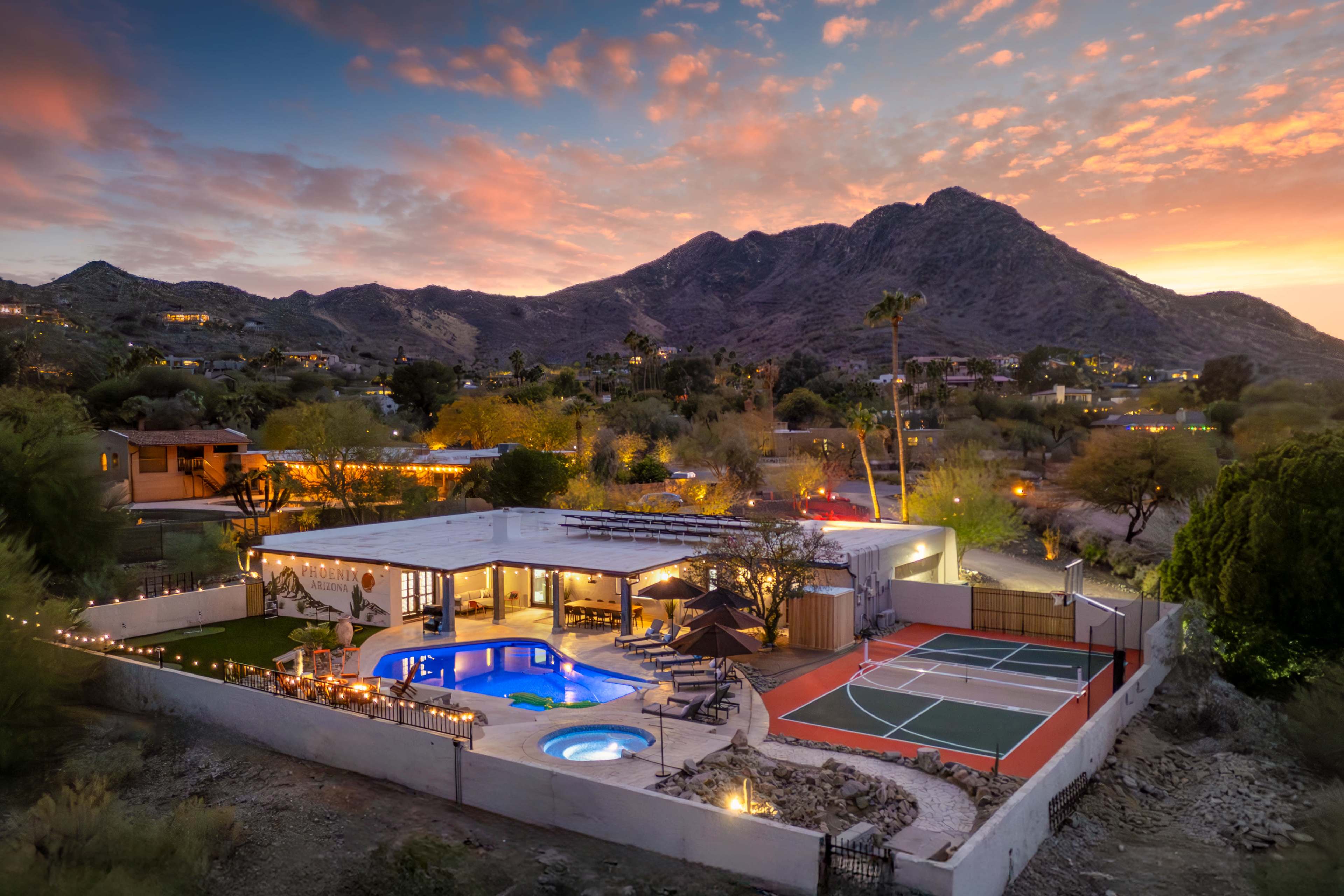 A modern home with a pool and tennis court is situated in a desert landscape, framed by mountains at sunset.