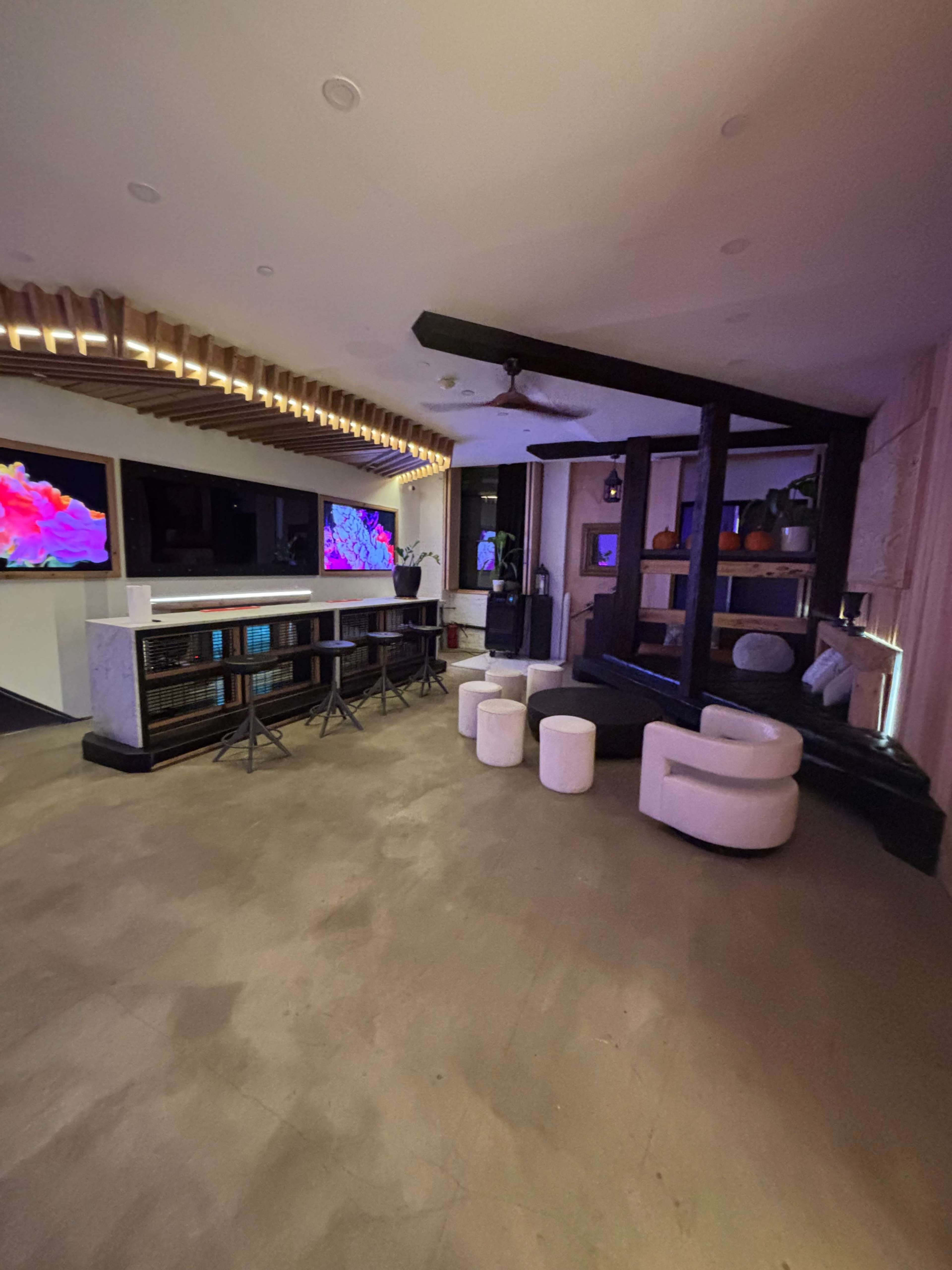 The image depicts a modern interior space featuring a bar area with stools, seating arrangements, and televisions mounted on the wall.