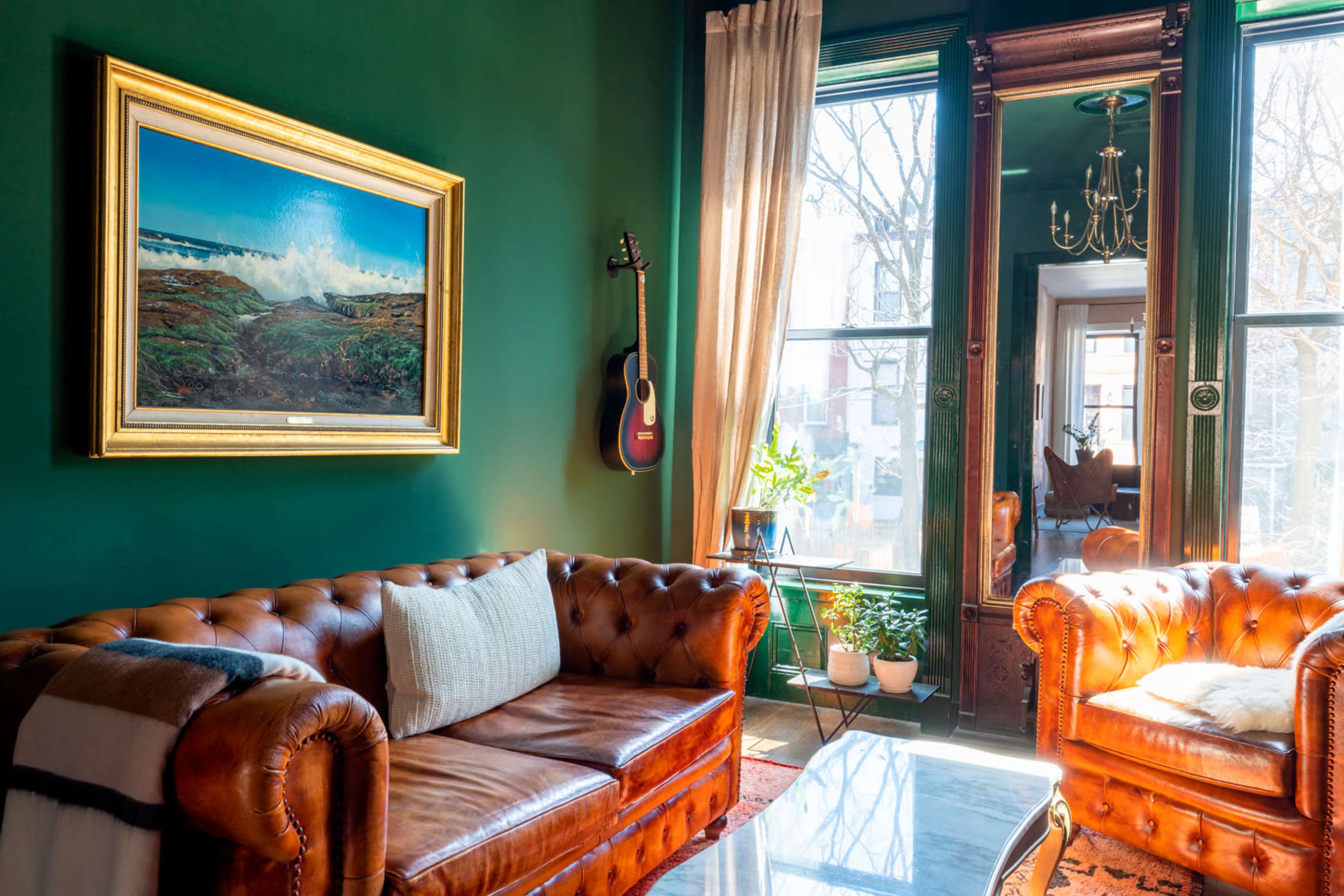 A cozy living room with dark green walls, a leather sofa, a marble coffee table, and a painting above the couch, alongside large windows bringing in natural light.