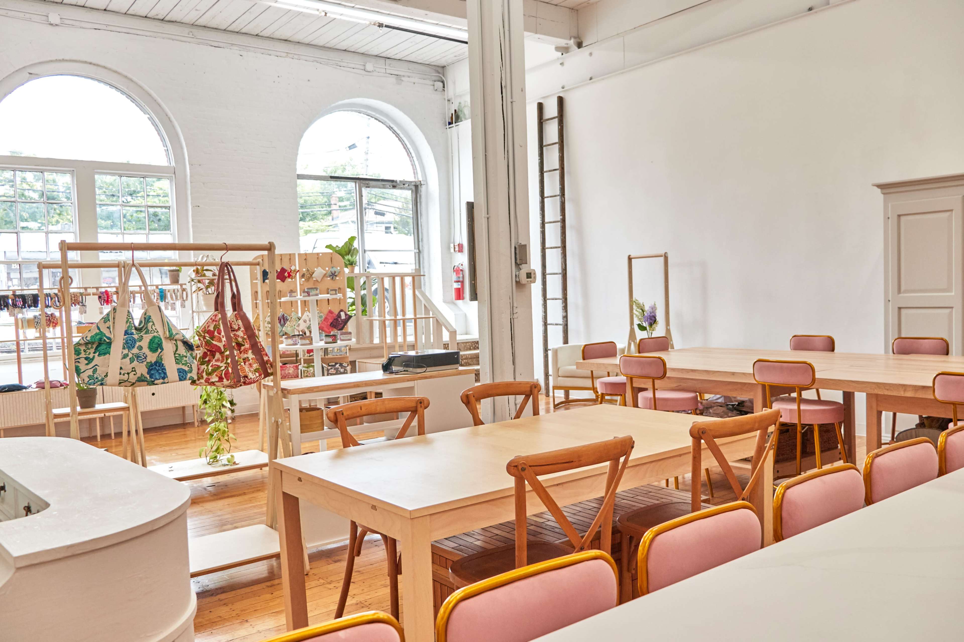 A spacious interior with wooden tables and pink chairs, featuring clothing displays and large arched windows.