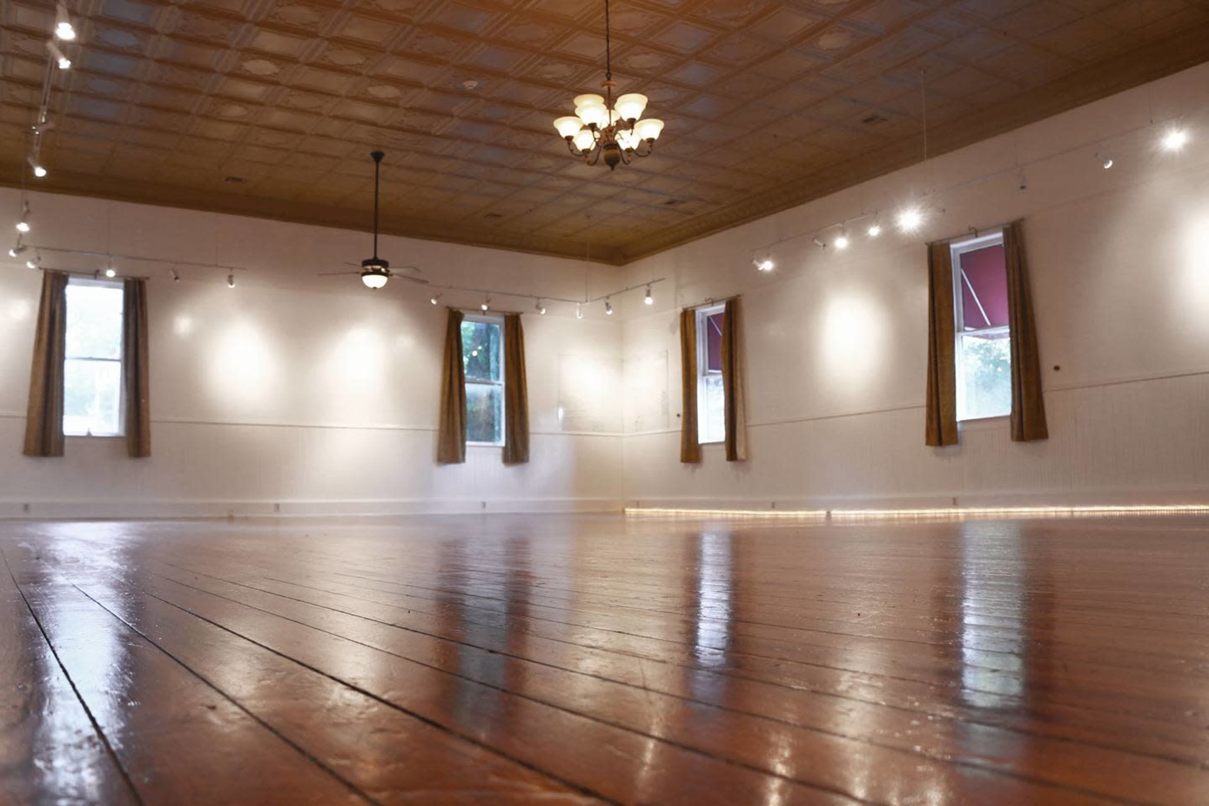 The image shows a spacious, empty room with polished wooden floors, large windows, and decorative lighting fixtures on the ceiling.