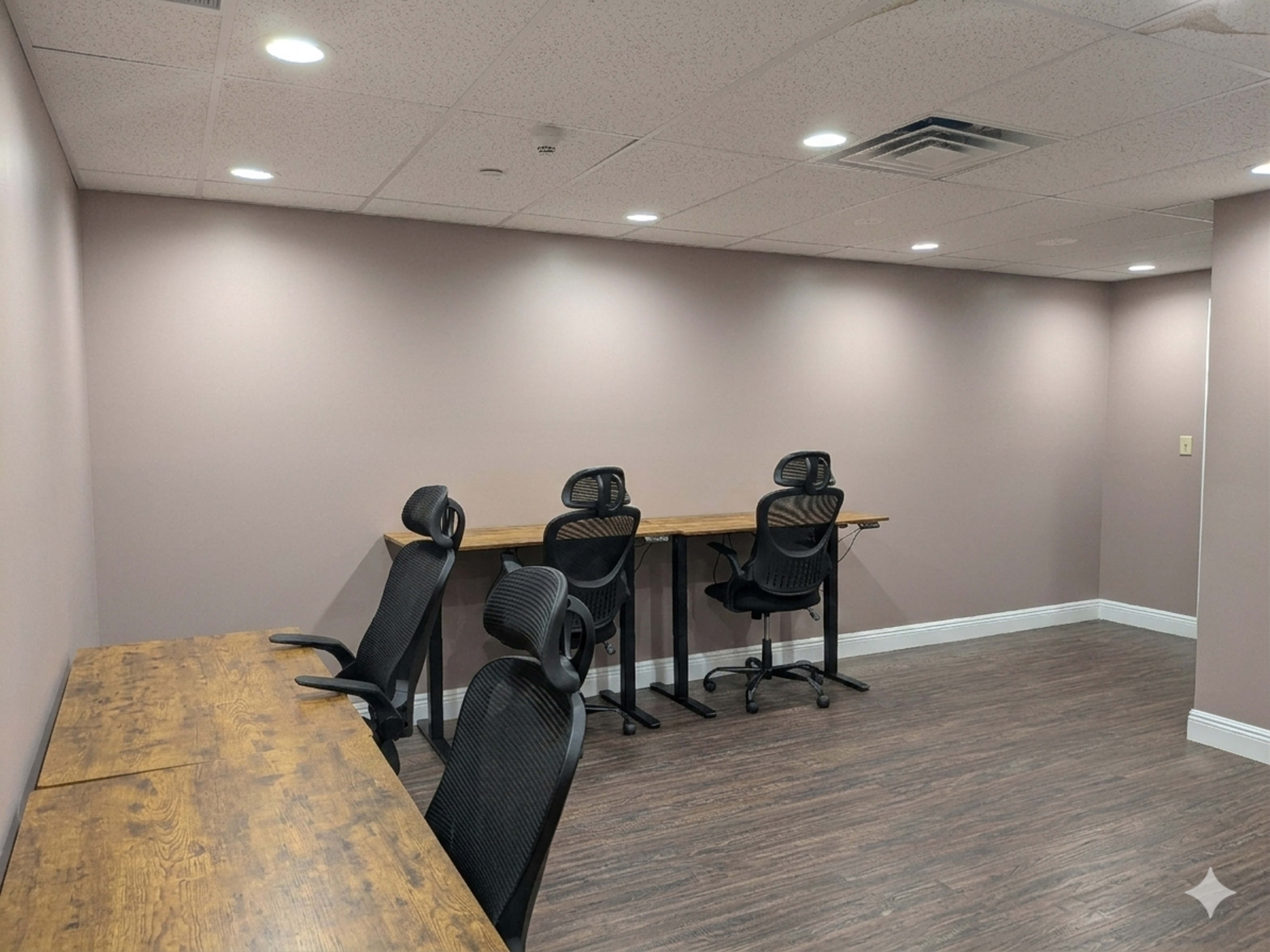 The image shows a minimalist office space with four black chairs facing two wooden desks against a plain pink wall.