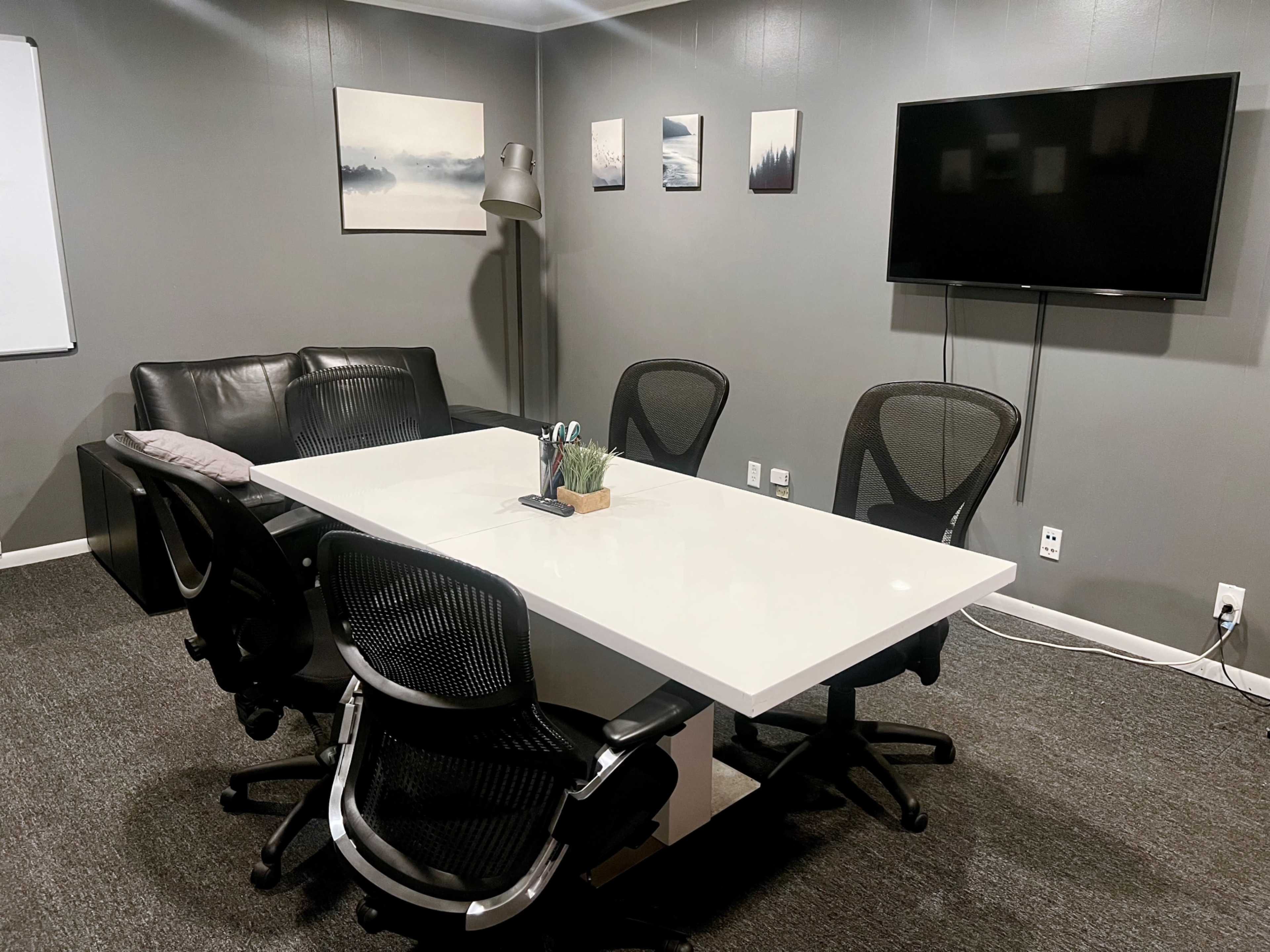 A modern conference room with a rectangular white table surrounded by four black office chairs, a sofa, a lamp, and a wall-mounted television.