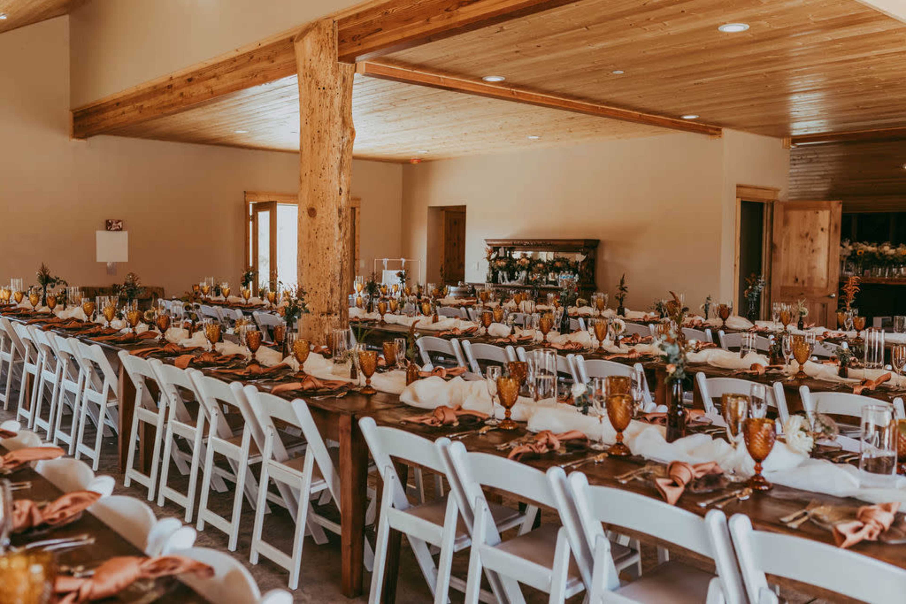 A spacious dining hall features long, elegantly set tables with white chairs, decorated with floral centerpieces and golden glasses.