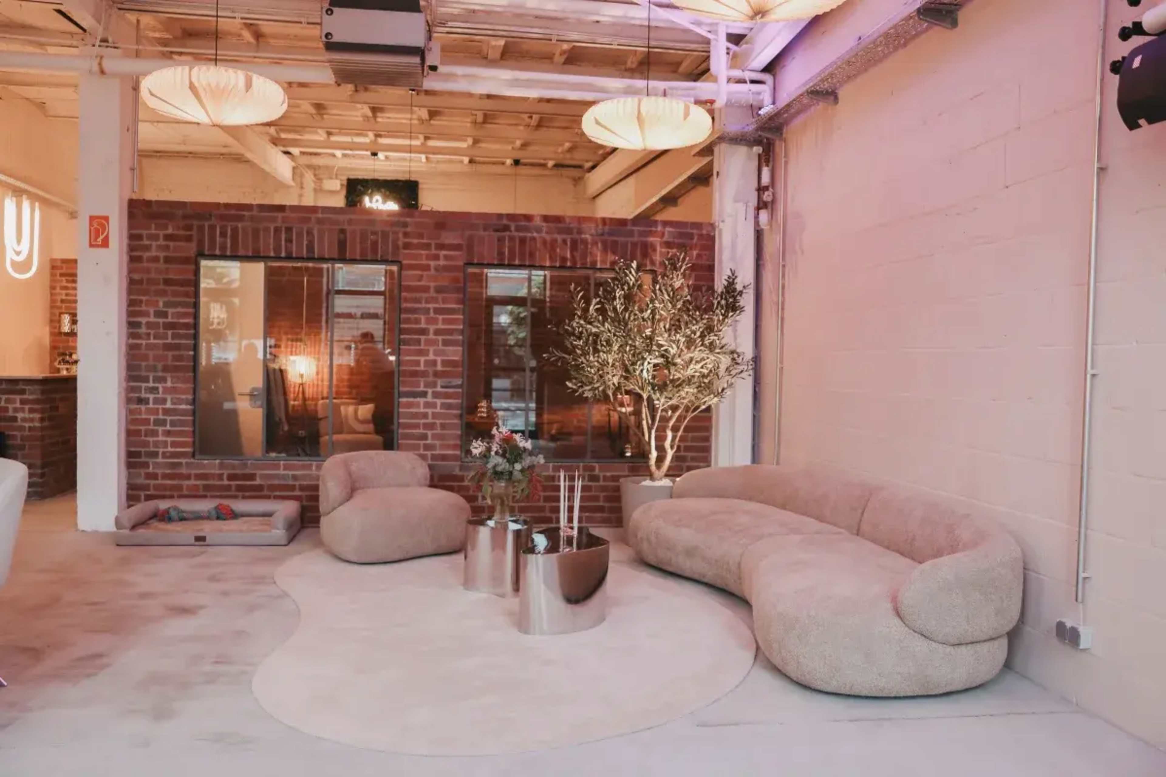 The image shows a modern lounge area featuring a curved beige sofa, a round rug, and a glass coffee table, with exposed brick walls and large windows in the background.