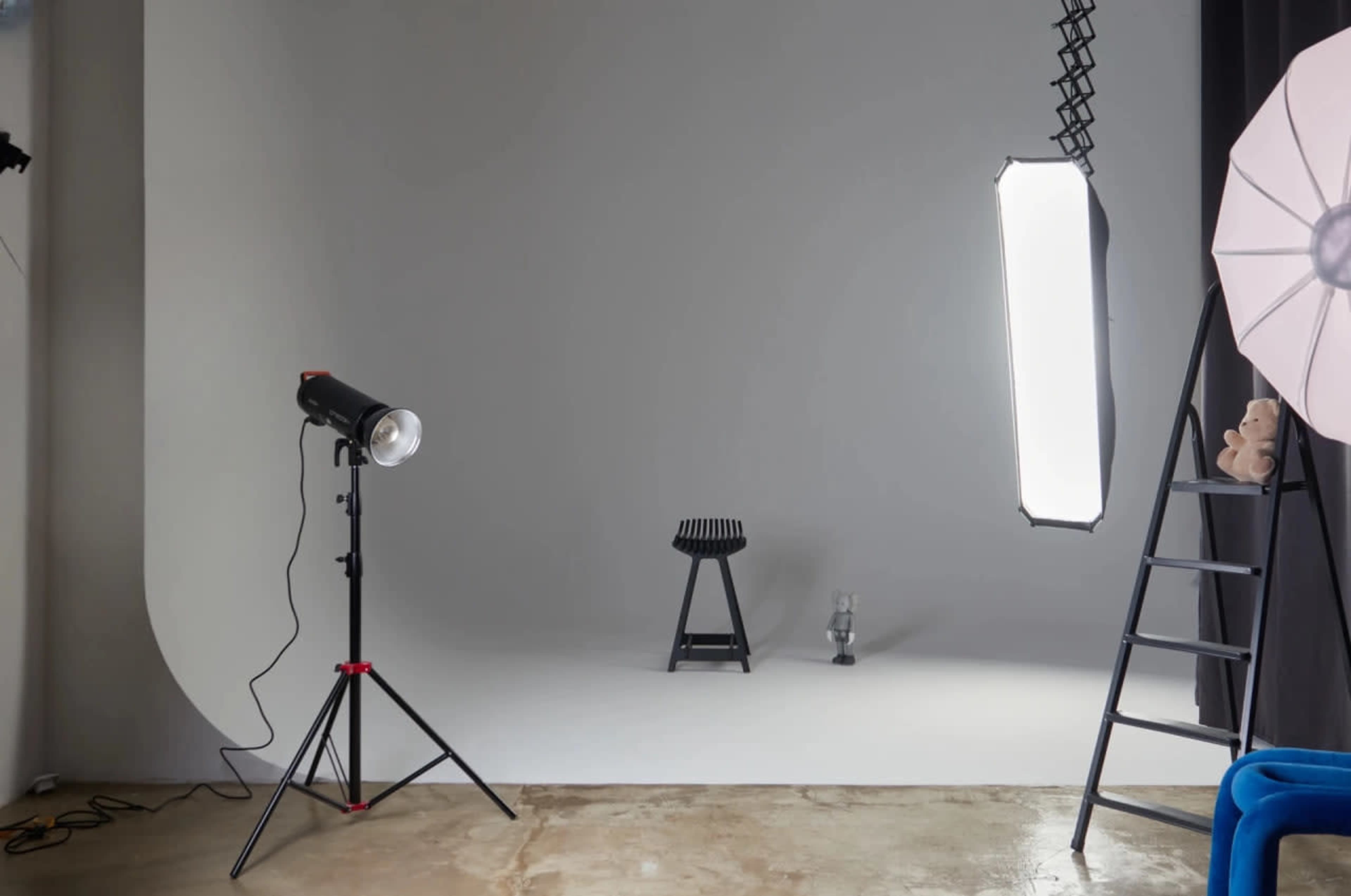 A photography studio is set up with a light source, a black stool, a glass vase, and a ladder against a plain gray backdrop.