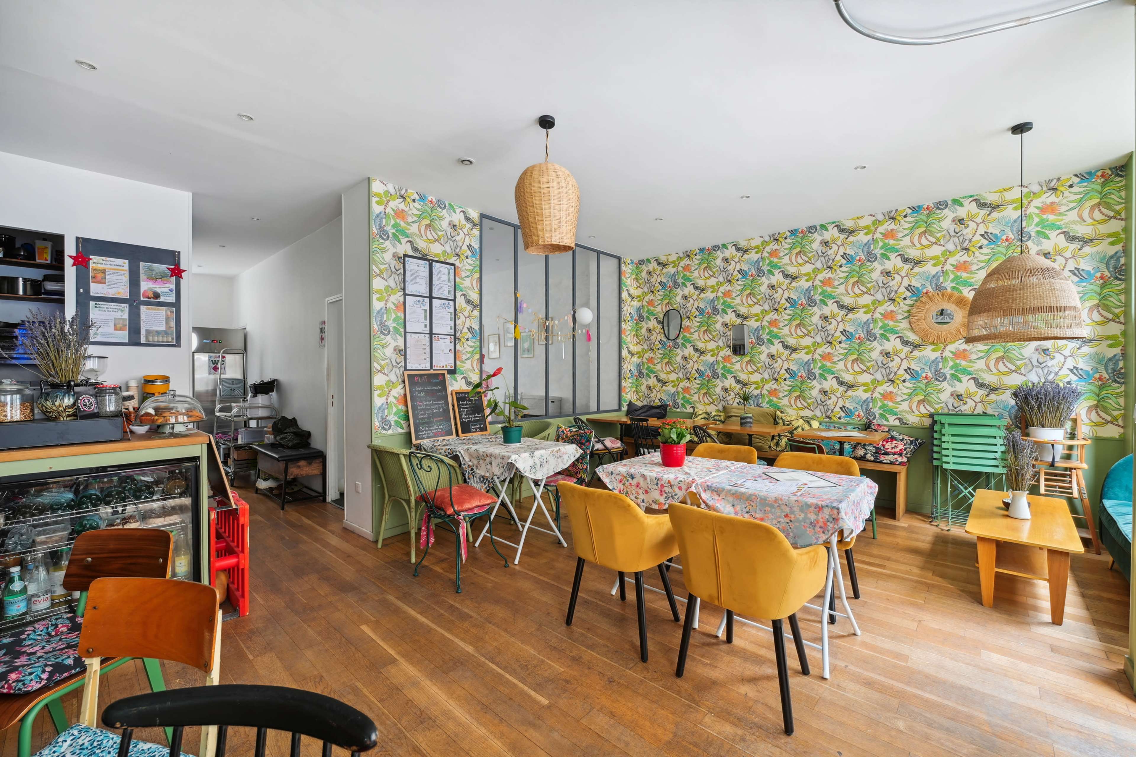 The cafe features colorful floral wallpaper, wooden flooring, and a mix of tables and chairs arranged for dining.