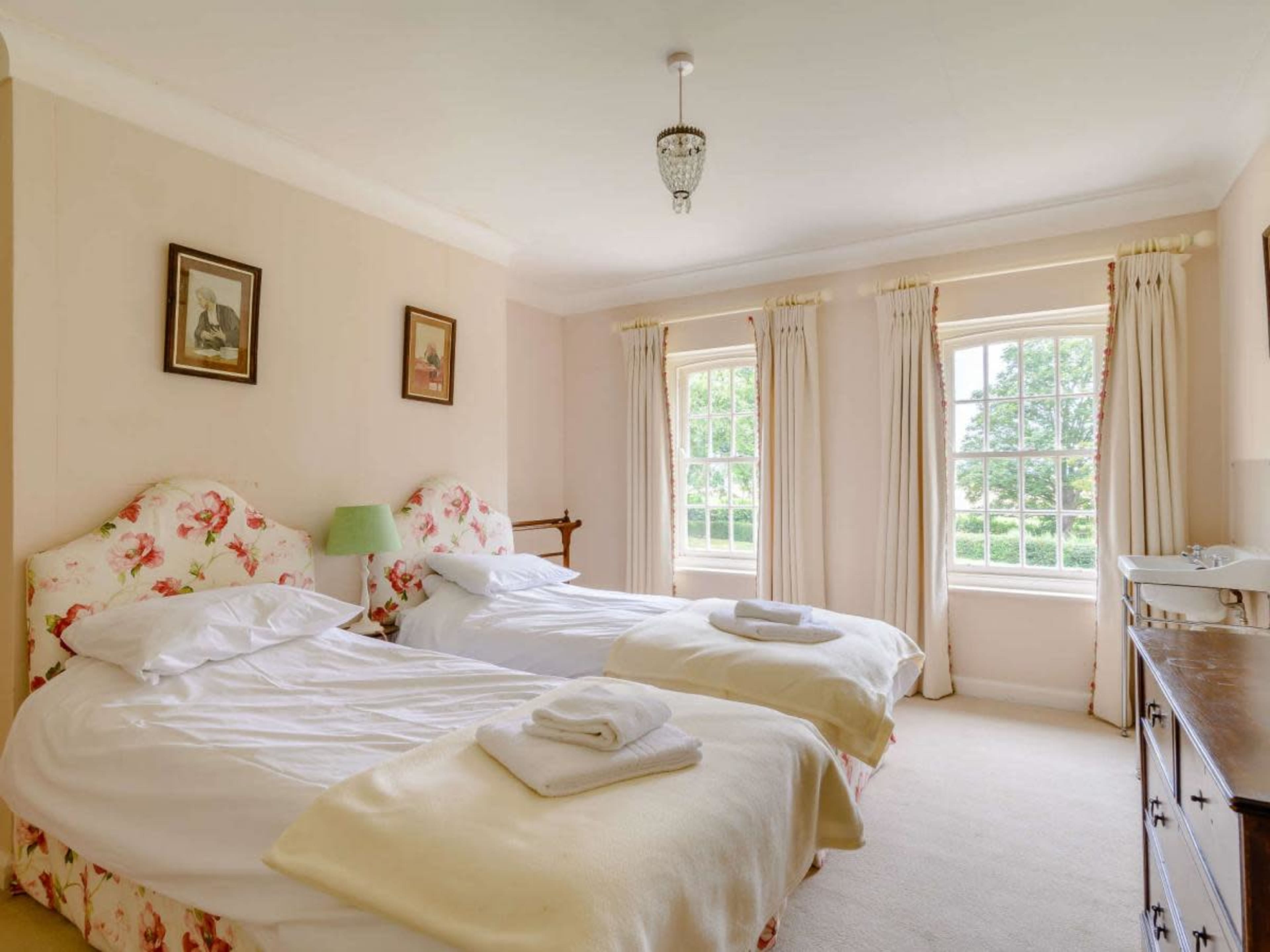 A well-lit bedroom featuring two twin beds with floral headboards, large windows with curtains, and a wooden dresser.