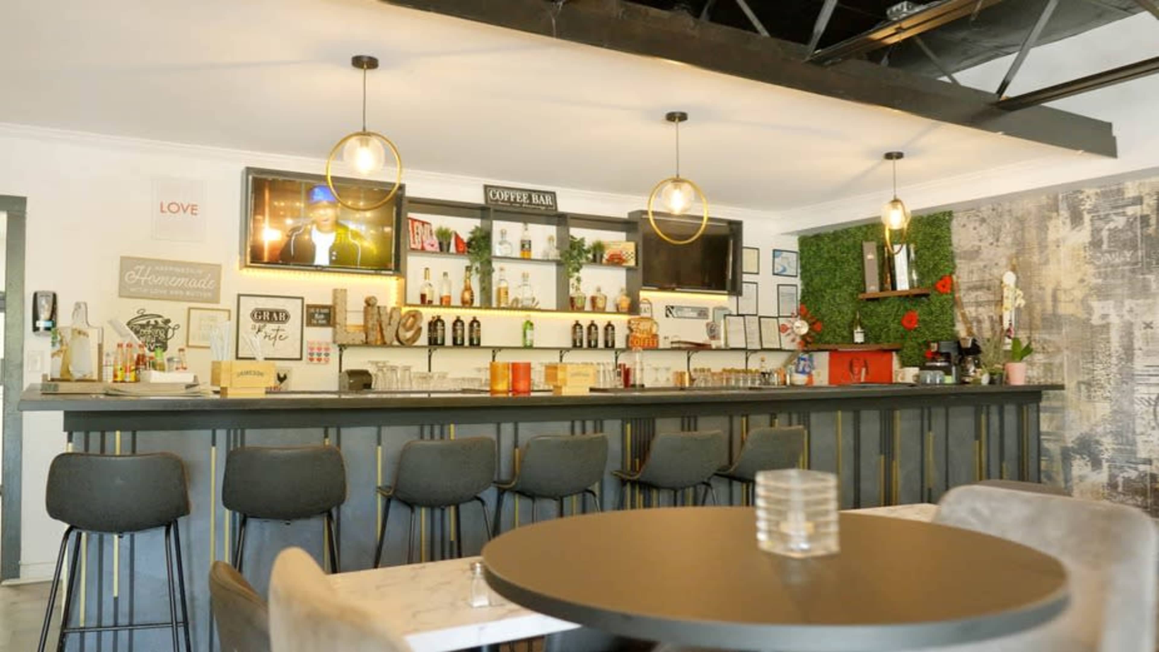 The image shows a modern bar area with high stools, a polished counter, shelves displaying various beverages, and decorative elements including plants and artwork.