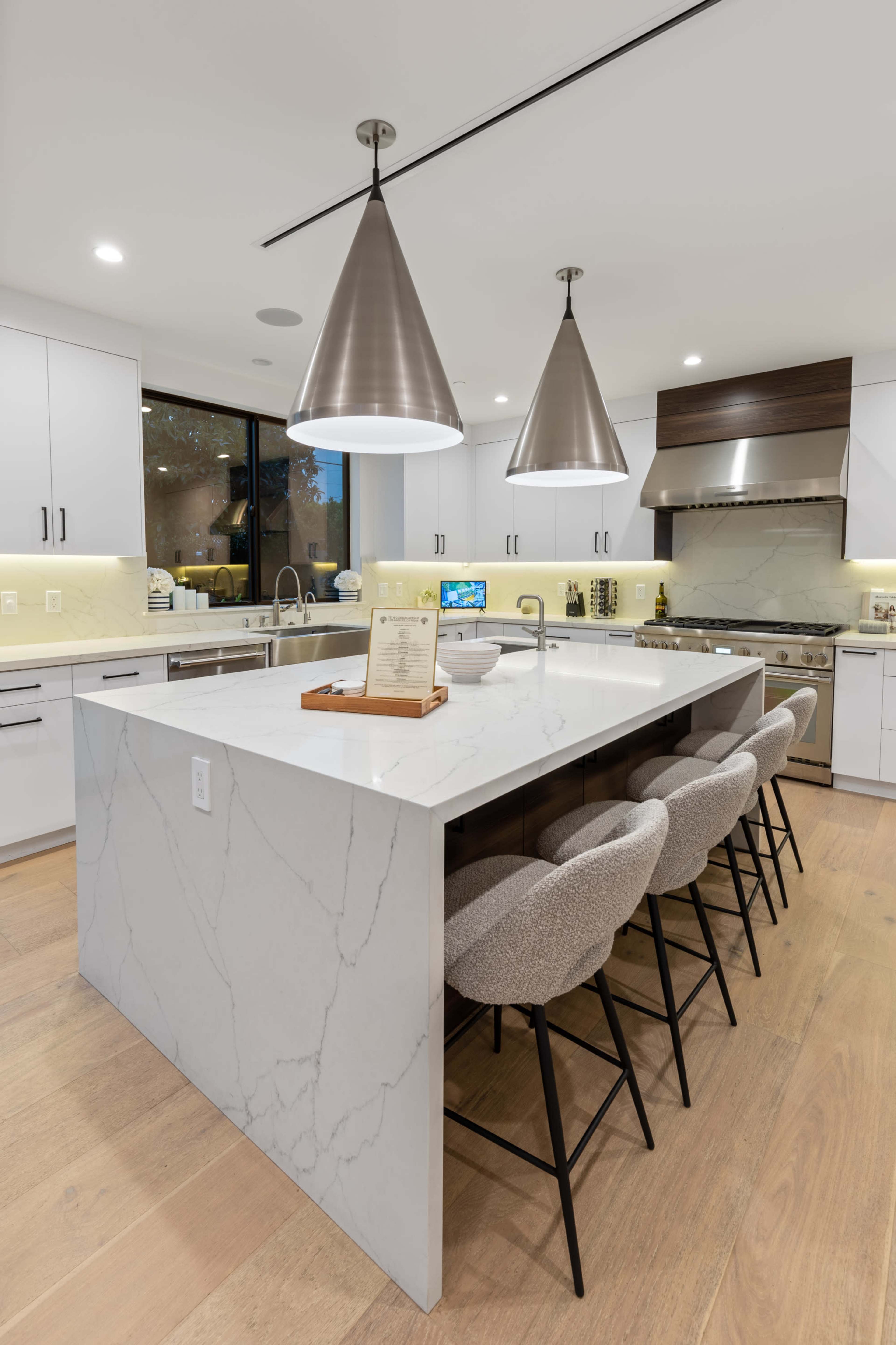 A modern kitchen features a large marble island with bar seating, sleek white cabinetry, and pendant lighting above.