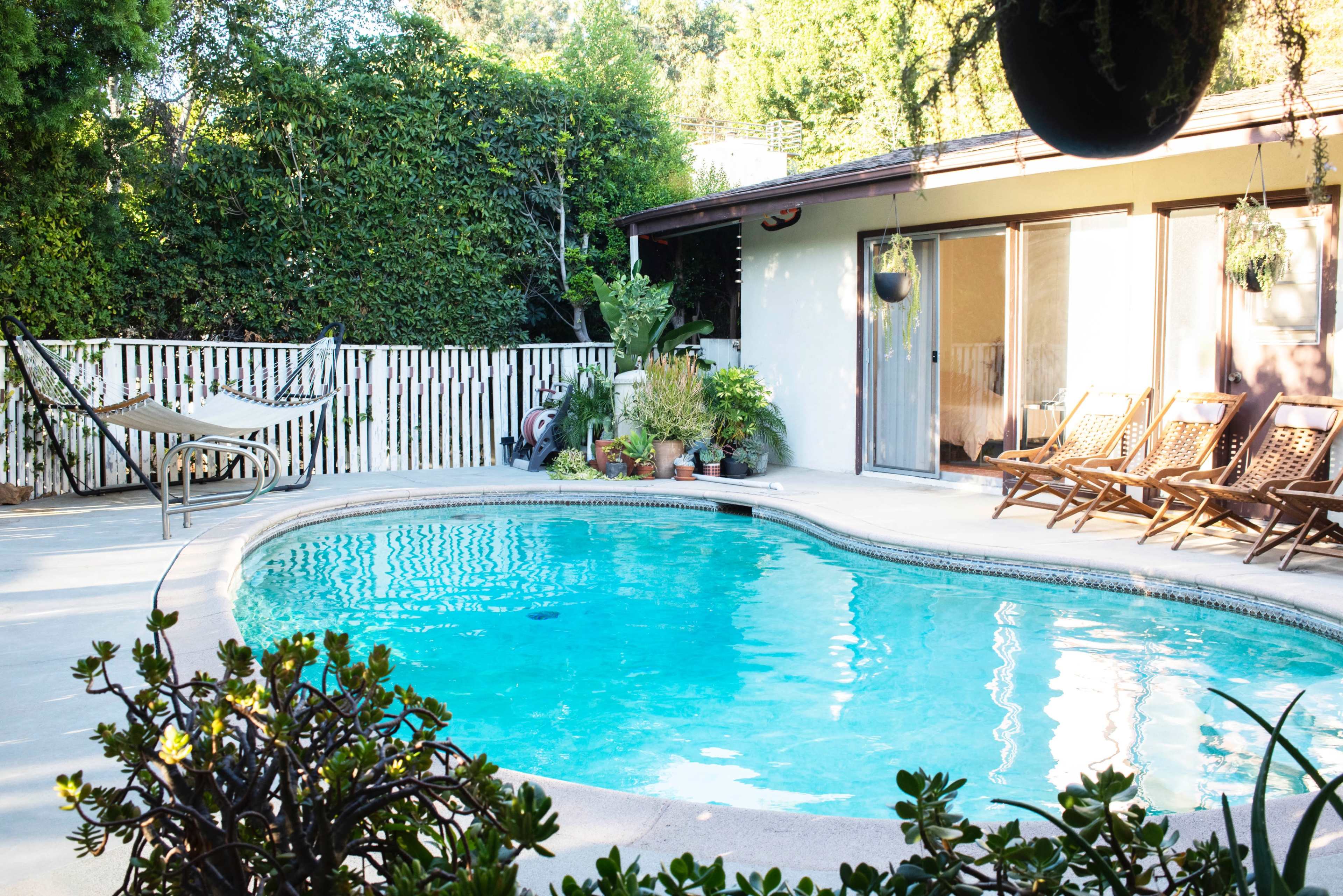 A backyard scene featuring a swimming pool surrounded by wooden lounge chairs, potted plants, and a hammock.