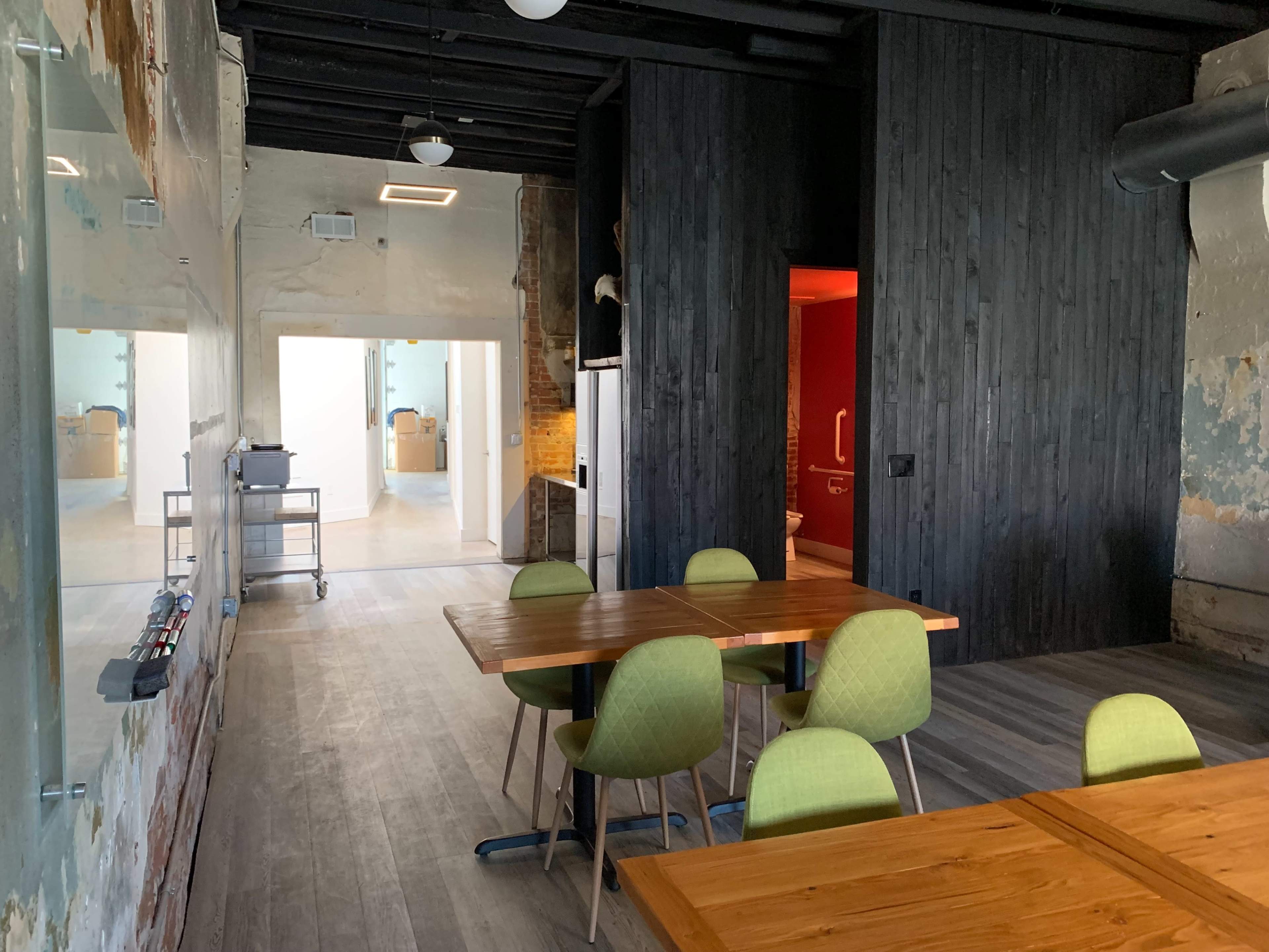A spacious, modern room features wooden tables and green chairs, with a view of a hallway leading to other rooms.