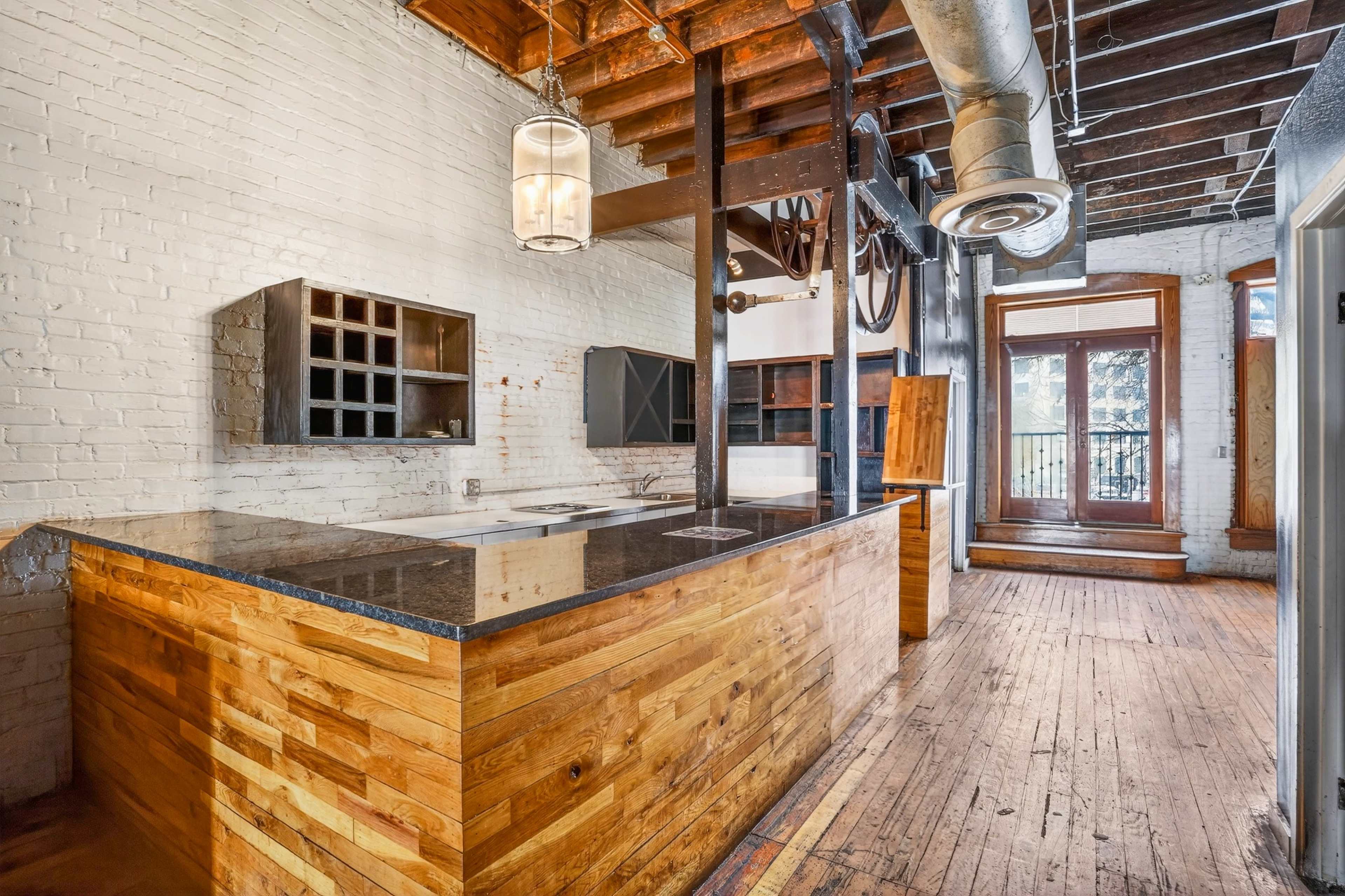The image shows a spacious kitchen with wooden countertops, exposed brick walls, and wooden floors, featuring an industrial design with hanging light fixtures and a visible old pulley system.