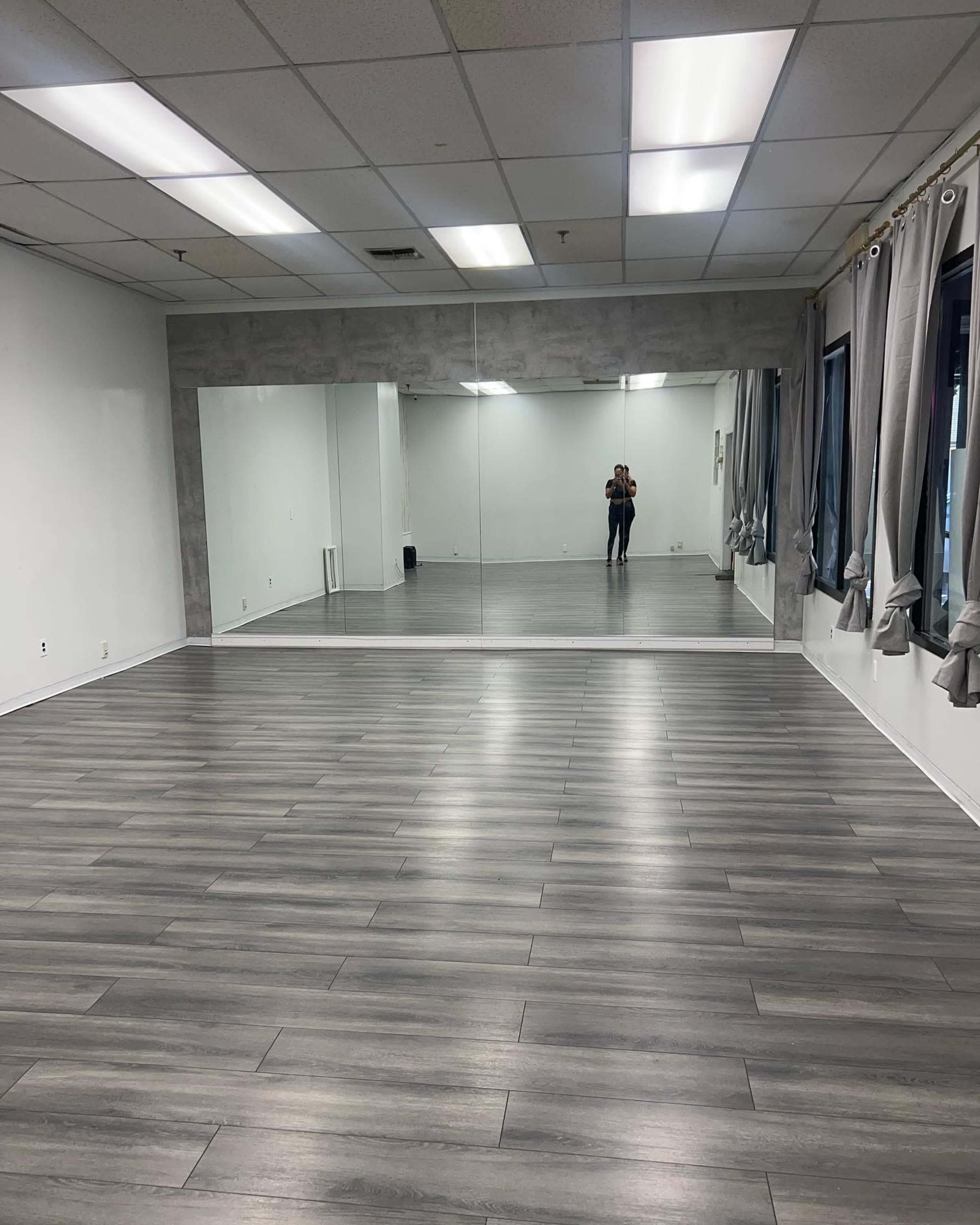 The image shows a spacious dance studio with wooden flooring, large mirrors on one wall, and gray curtains at the windows.