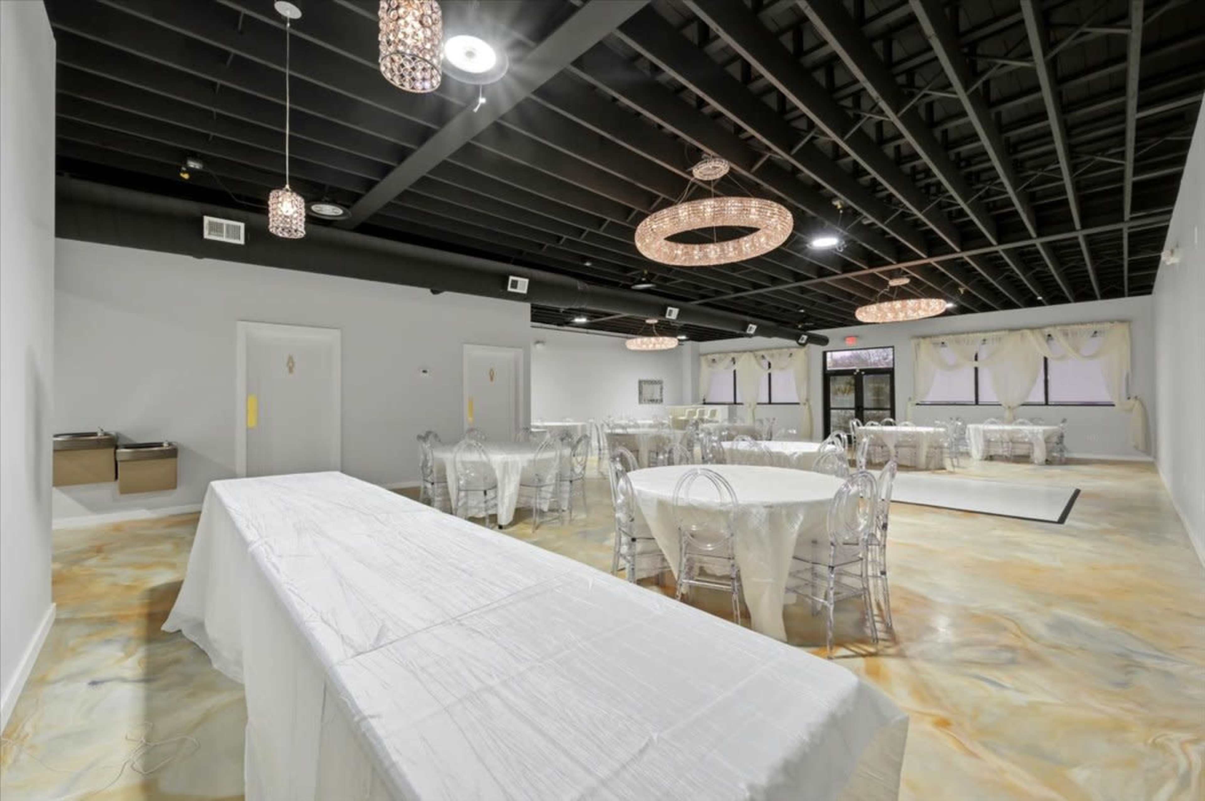 The image shows a spacious, well-lit event room with round and rectangular tables set with white linens and clear chairs, along with decorative lighting fixtures hanging from the ceiling.