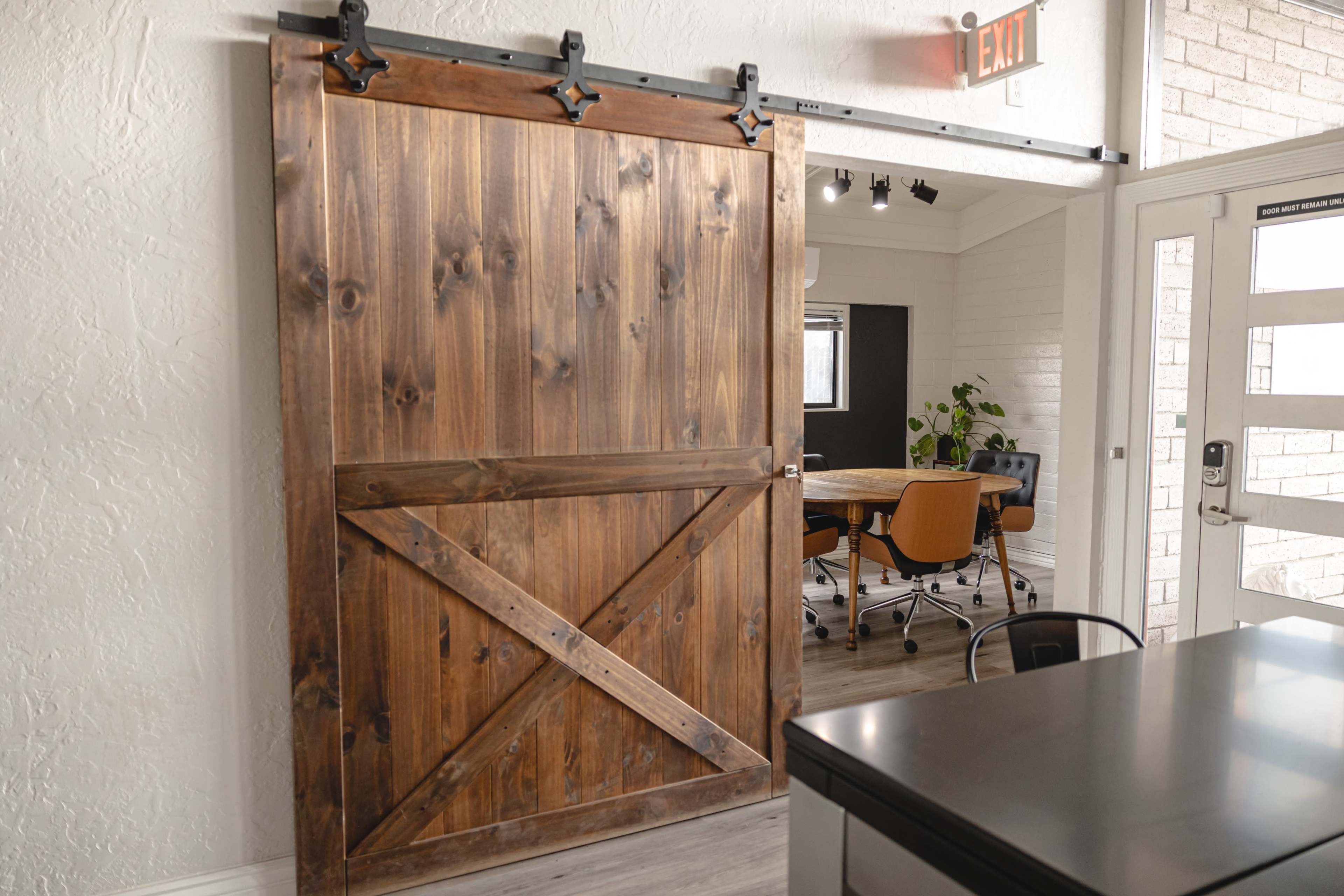 A wooden barn door with a sliding track opens to a modern conference room featuring a round table and orange chairs.