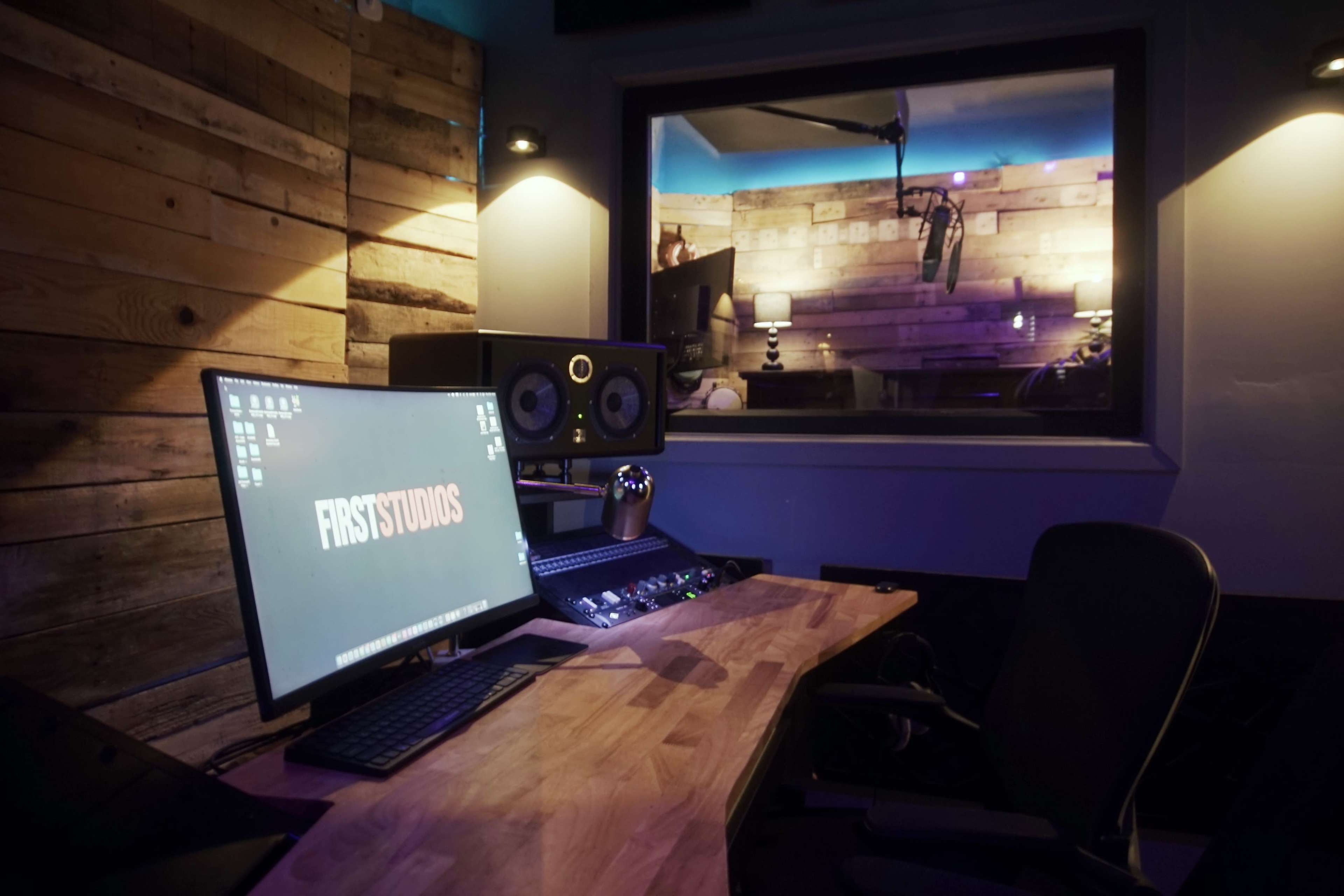 The image shows a modern music production studio with a computer workstation featuring a large display screen and audio equipment, adjacent to a soundproof recording booth.