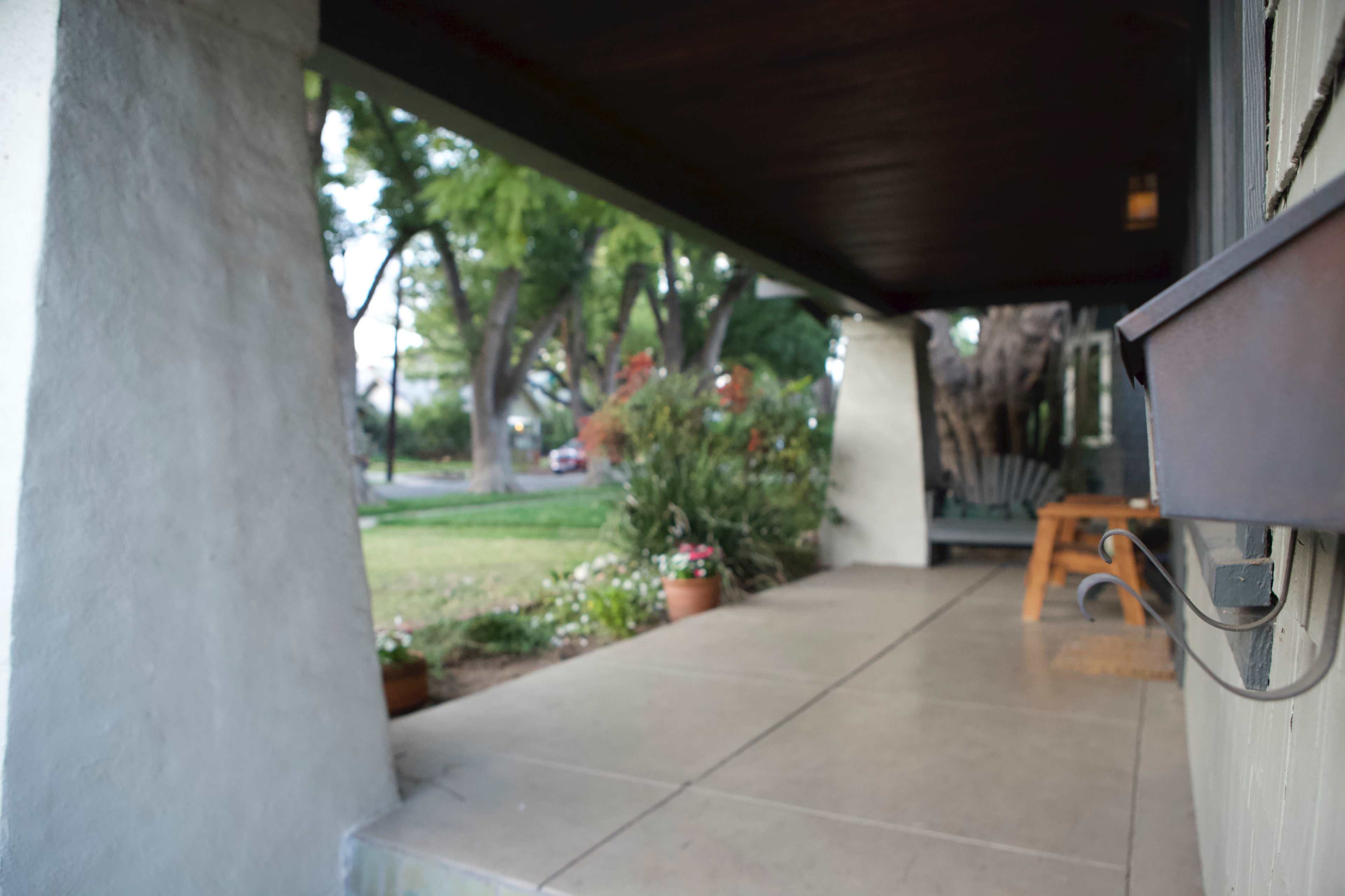The image shows a covered porch with concrete flooring, flanked by two columns, and plants in pots alongside a view of the yard and street.