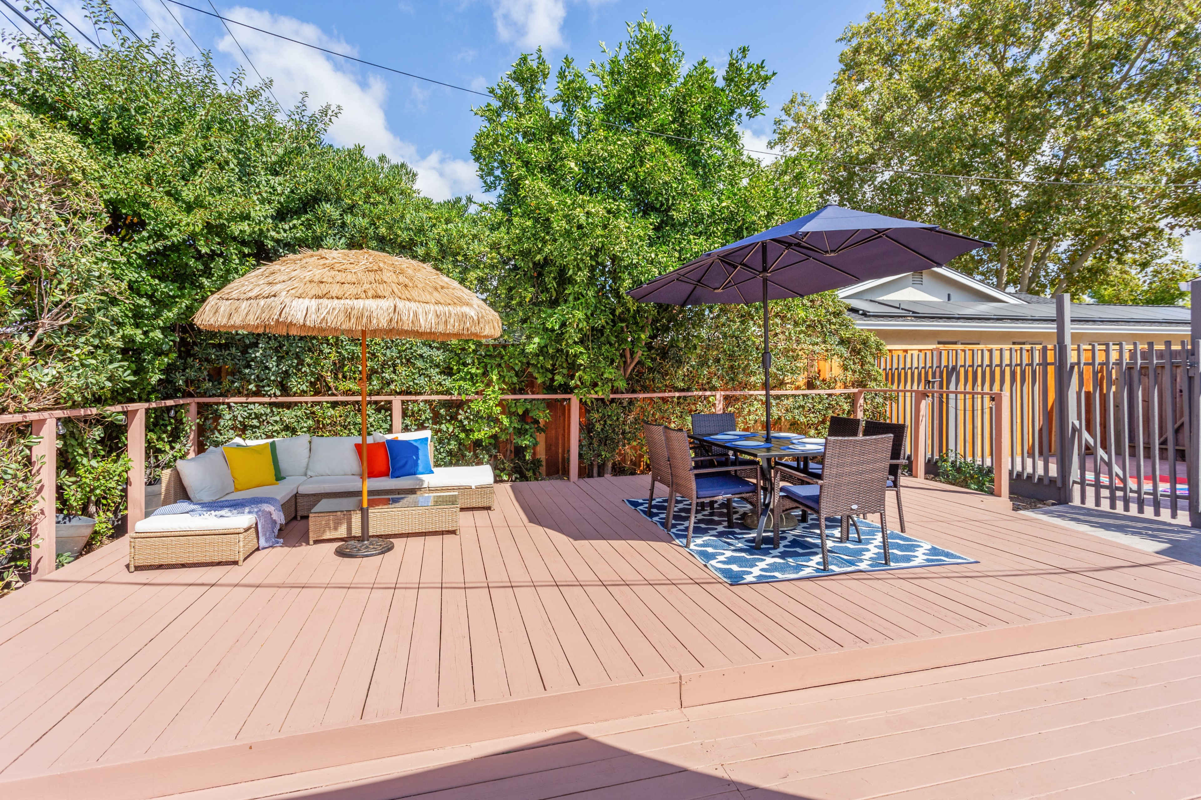 A wooden deck featuring a shaded seating area with a thatched umbrella, a dining table set beneath a black umbrella, and vibrant greenery in the background.