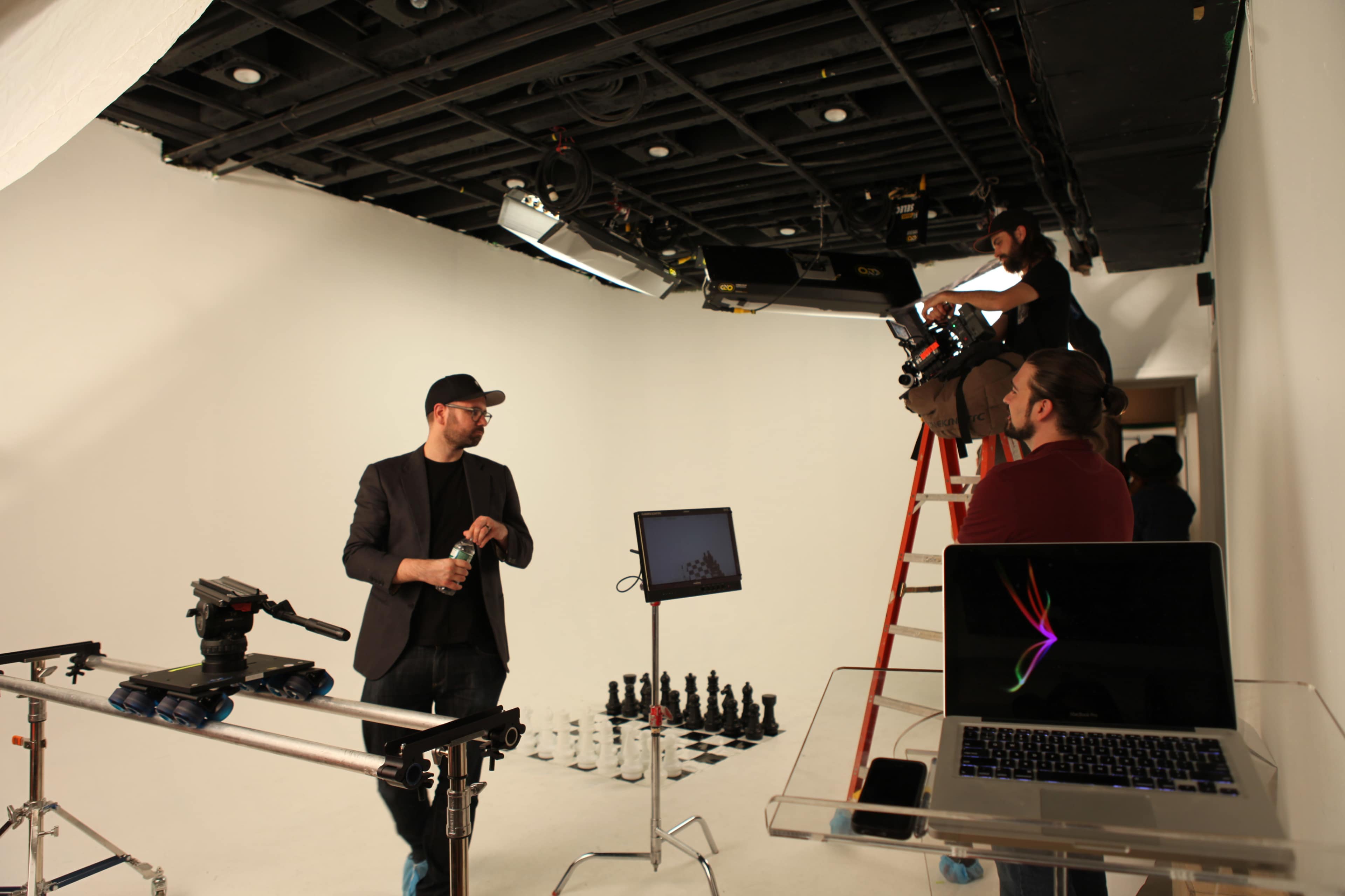 A film crew is preparing a set in a studio with a white backdrop, featuring a chessboard and equipment including cameras and a laptop.