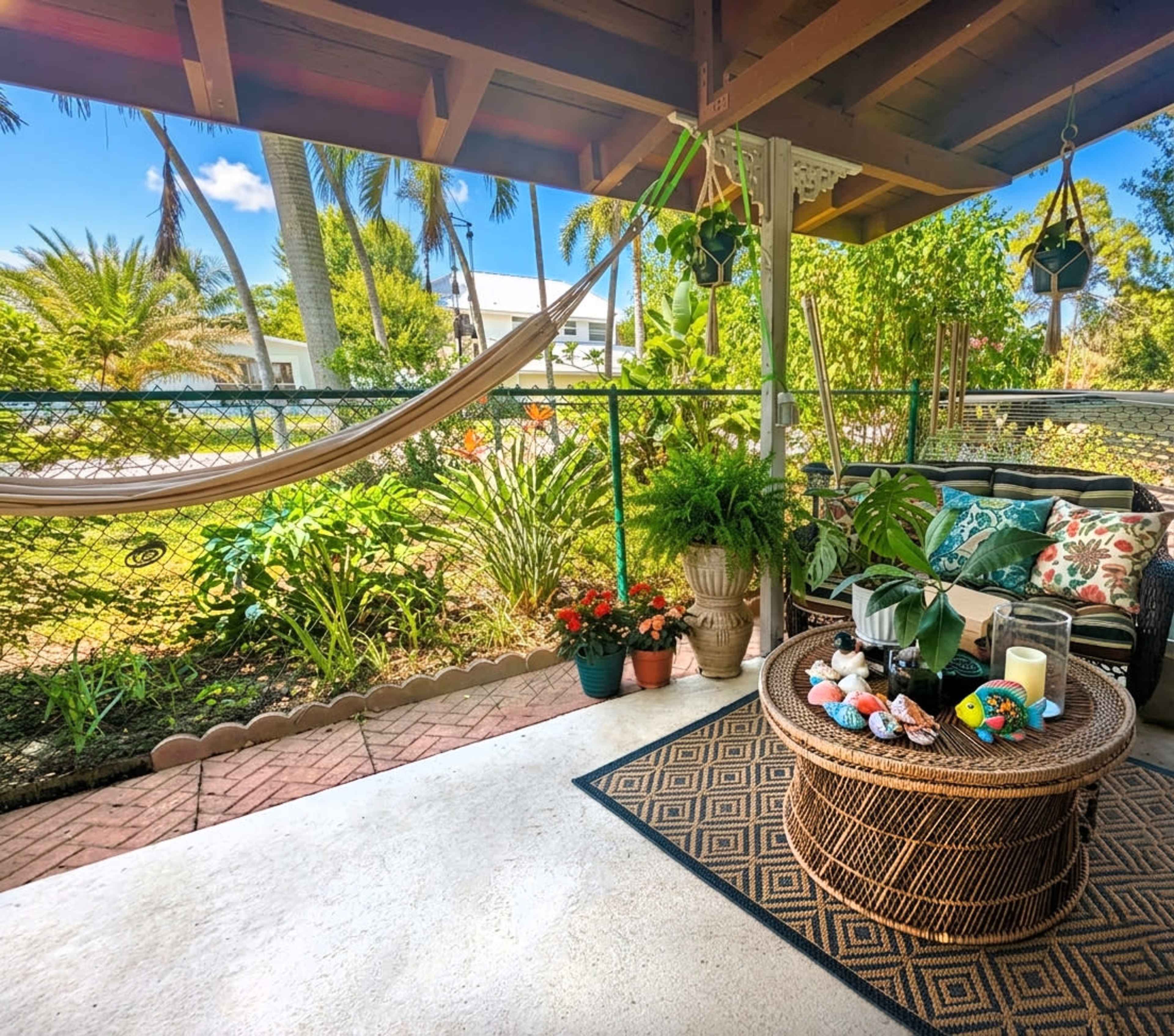 A porch features a round wicker coffee table surrounded by plants, cushions, and a hammock, with a view of a garden and blue sky in the background.