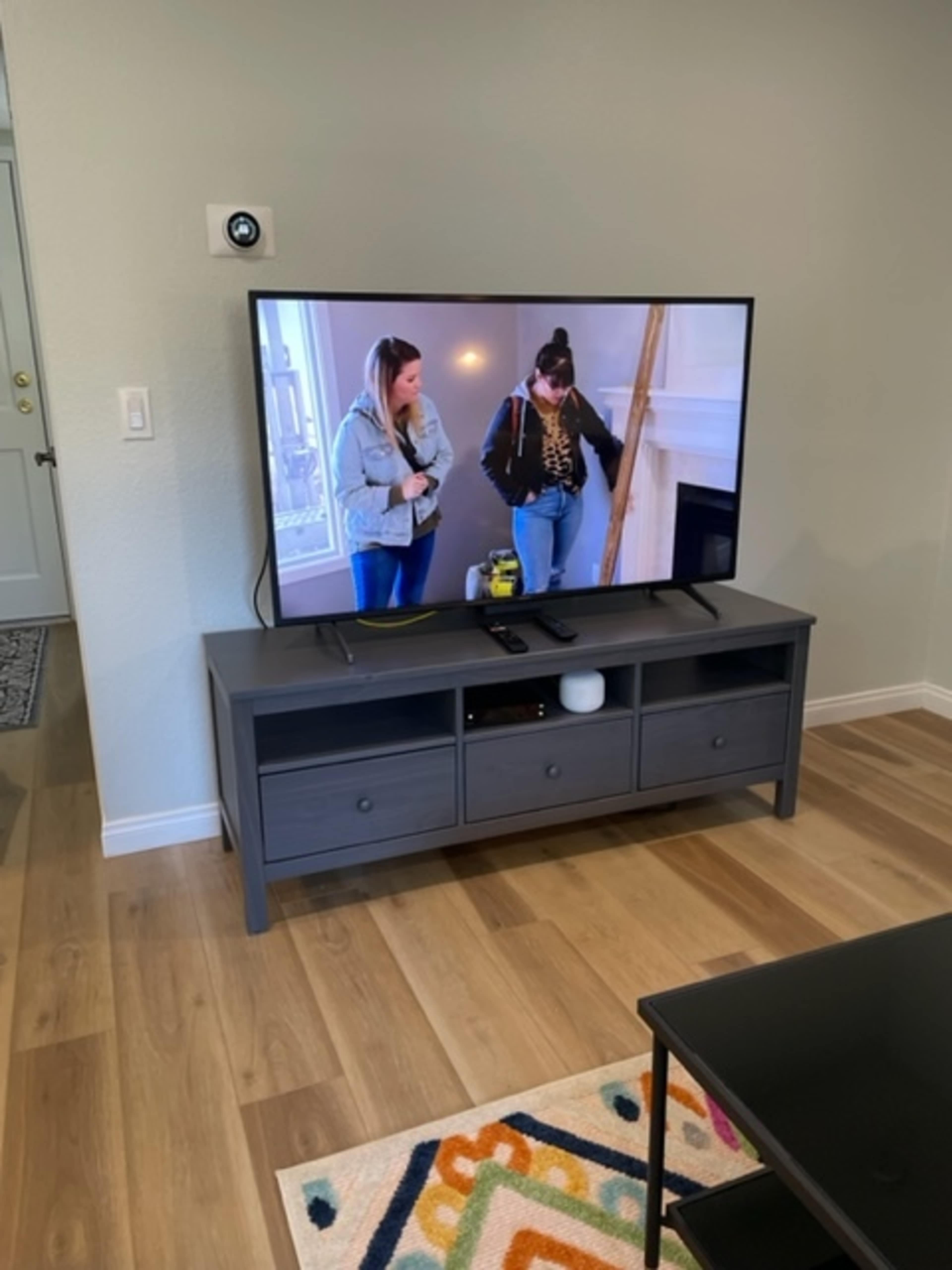 A television displaying a scene with two women is placed on a gray media console in a living room with wooden flooring.