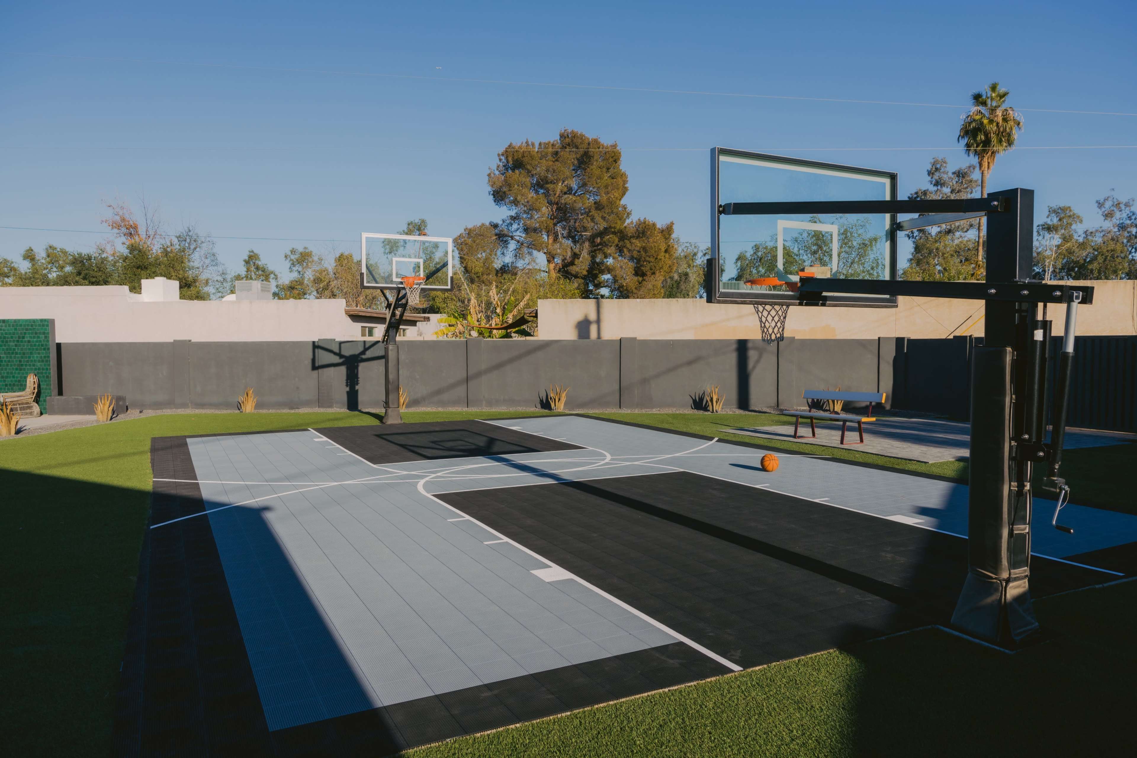 A basketball court with two hoops, surrounded by greenery and a clear blue sky.