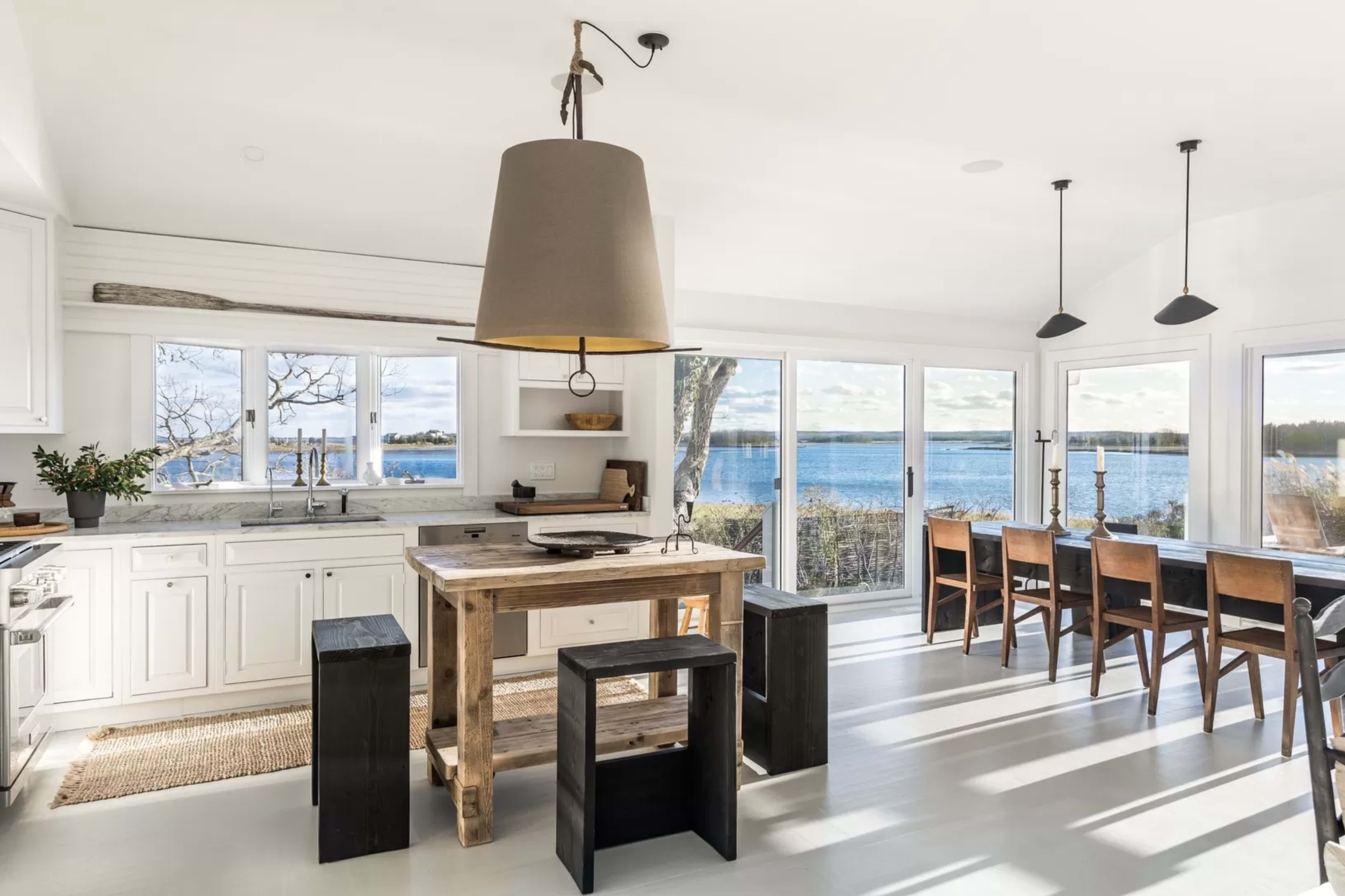 A bright kitchen features a wooden island with stools, large windows showcasing a view of the water, and a dining area with a table and chairs.