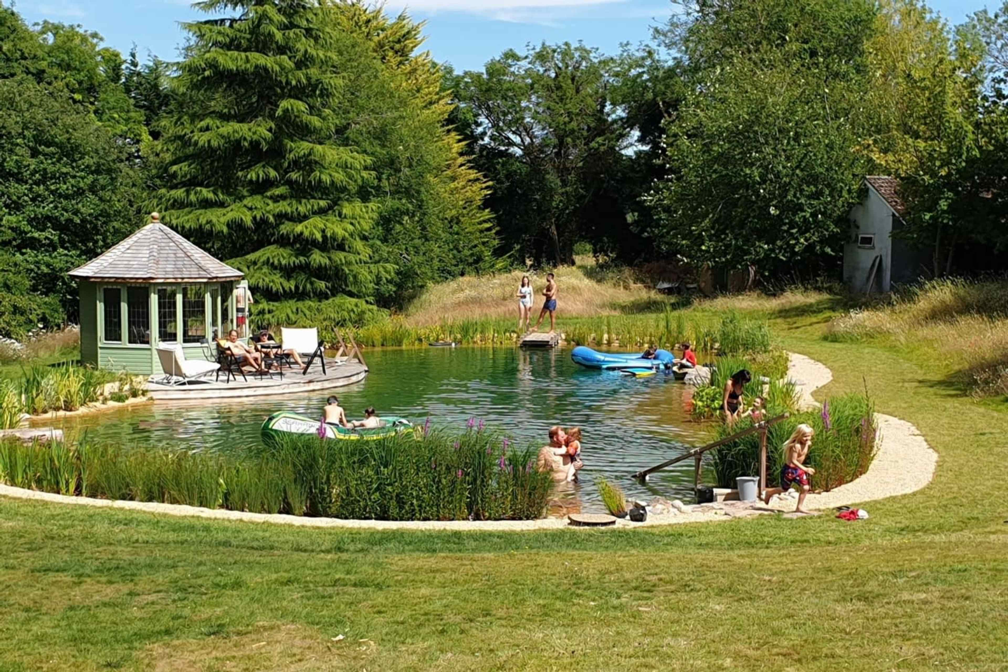 People enjoy a sunny day by a natural swimming pond surrounded by greenery and a gazebo.