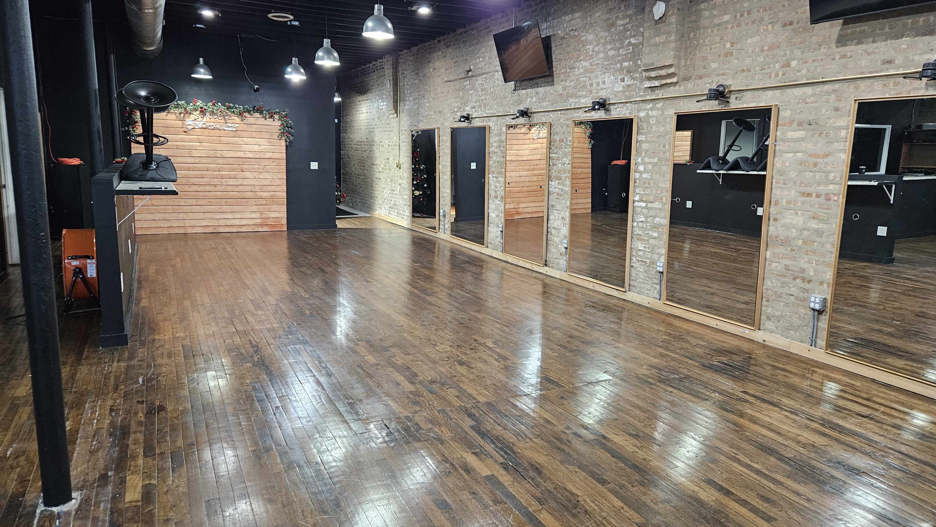 The image shows an empty studio space with polished wooden floors, mirrored walls, and black accents.