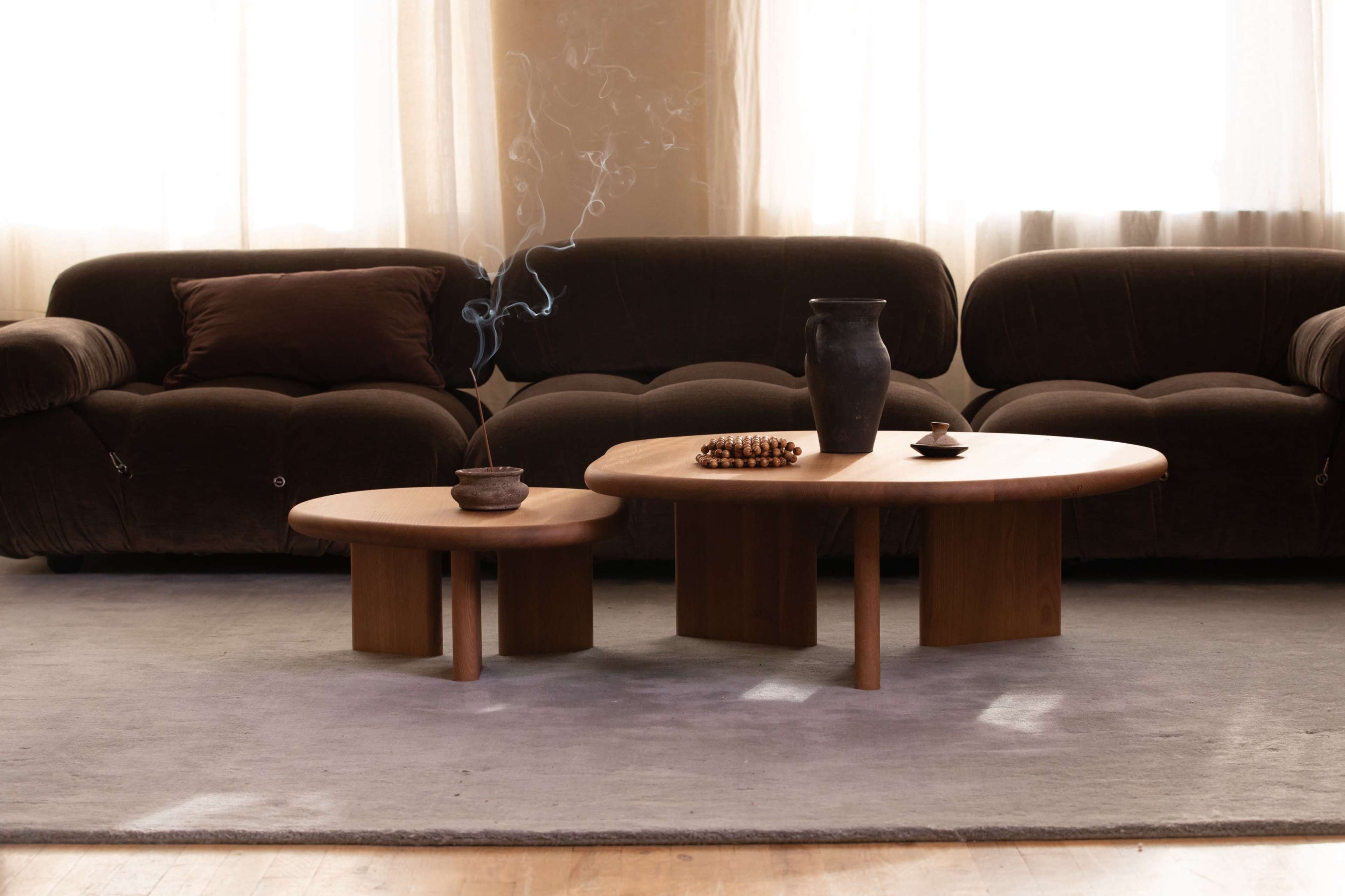 A living room scene features two wooden coffee tables, one smaller and round, placed in front of a brown velvet sofa, with a ceramic vase and a bowl on the tables, and smoke curling upward from a stick incense.