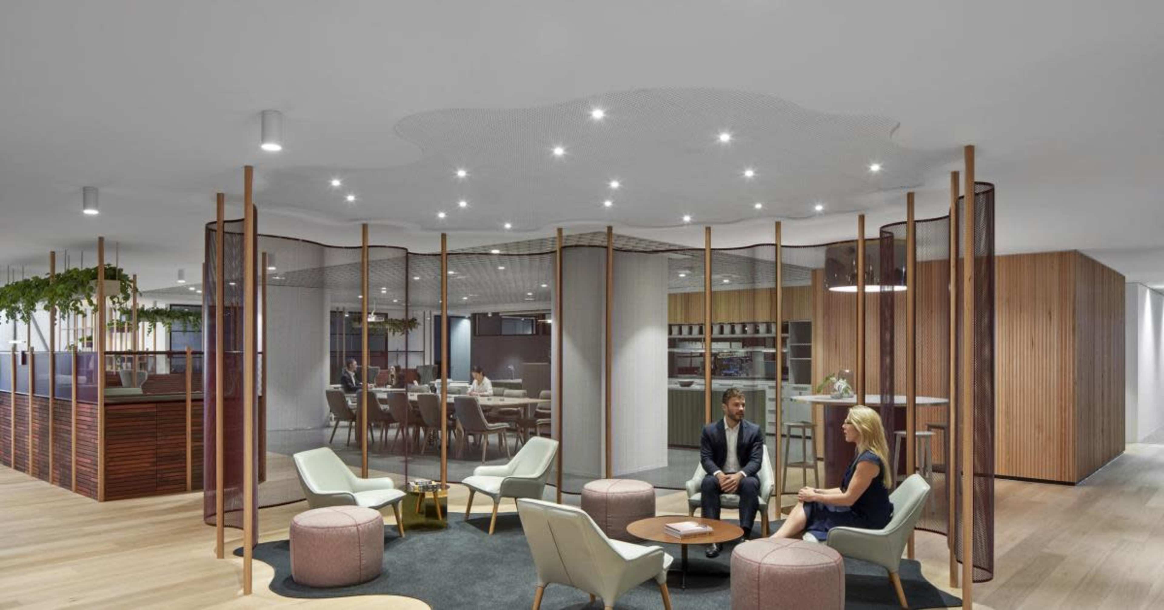 The image depicts a modern office lounge area with two people seated on a circular rug, surrounded by contemporary furniture and glass partitions.