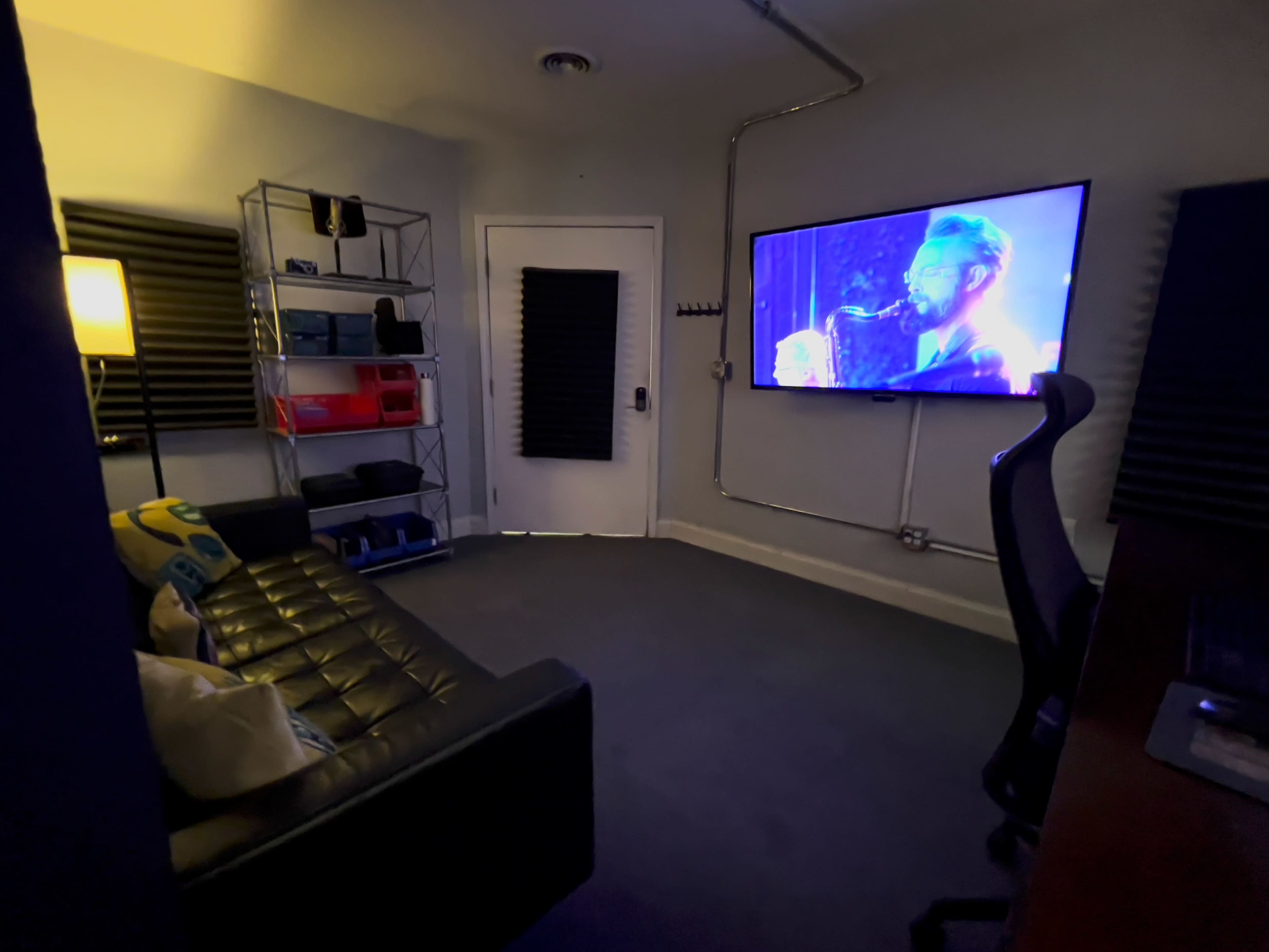 The image shows a small room with a black couch, a large wall-mounted television displaying a performance, and a shelf containing various items.