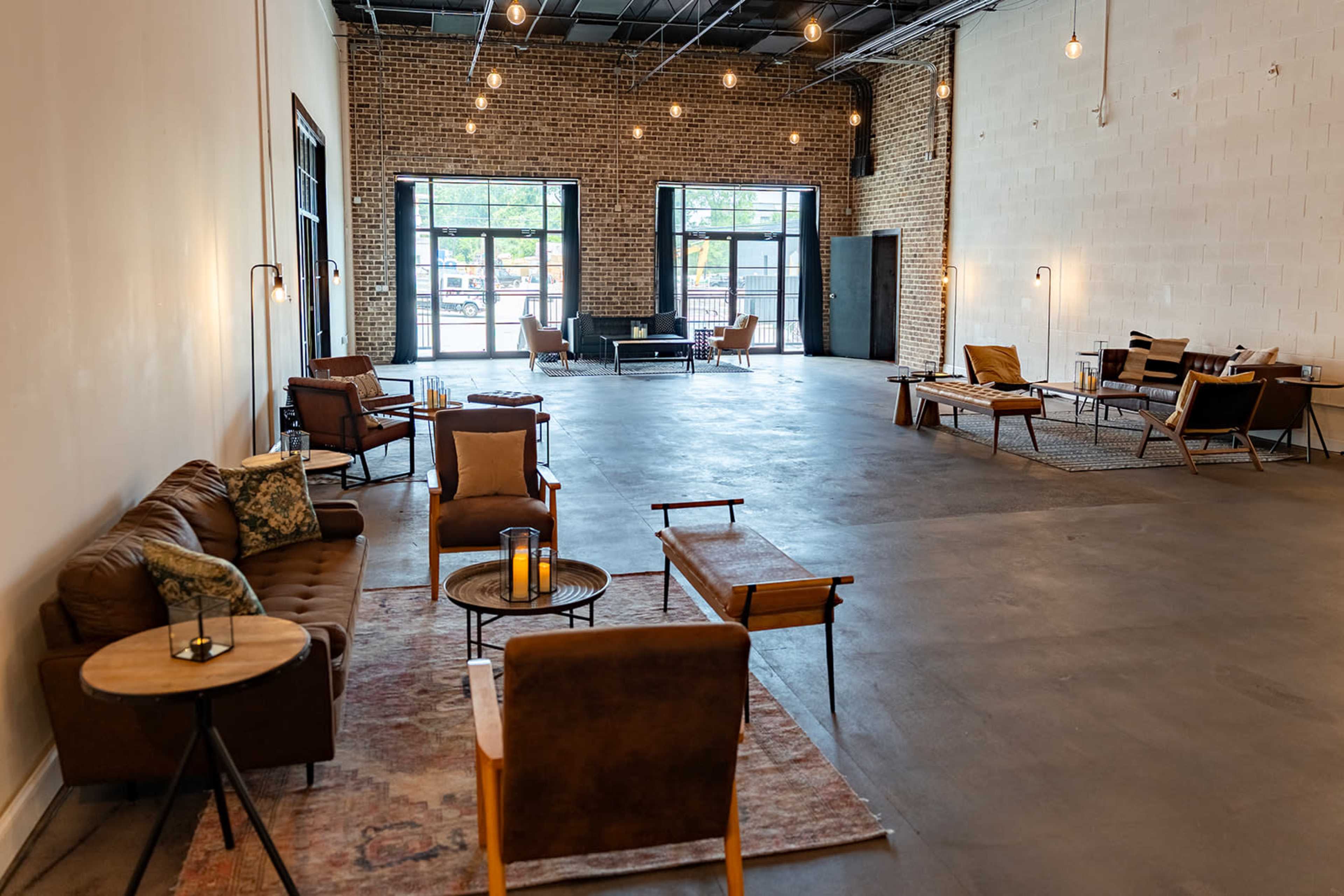 The image shows a spacious, well-lit room with modern seating arrangements, large windows, and an industrial brick wall.