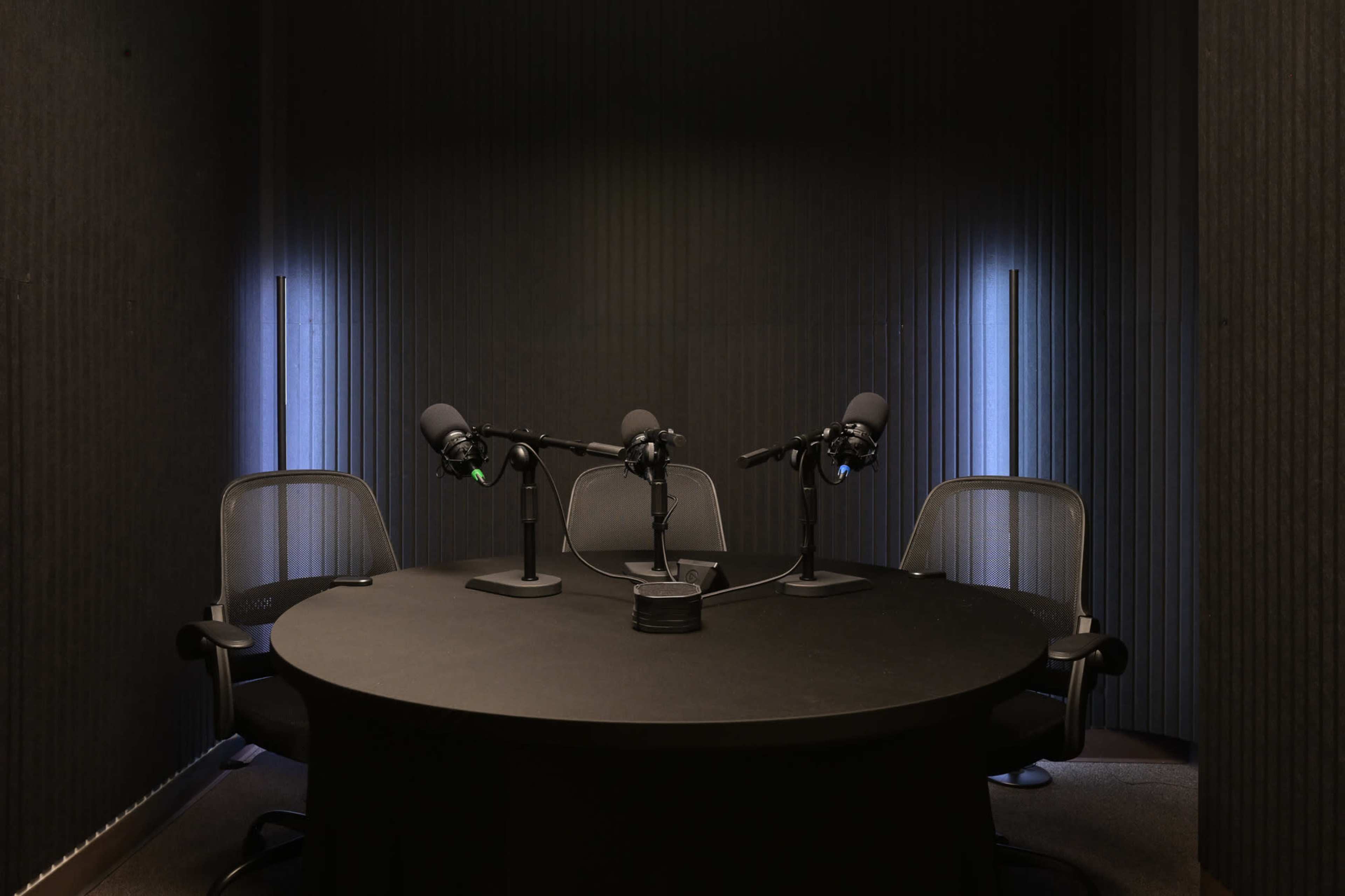 A soundproof recording studio features a round table with three microphones and two chairs, all surrounded by dark acoustic panels.