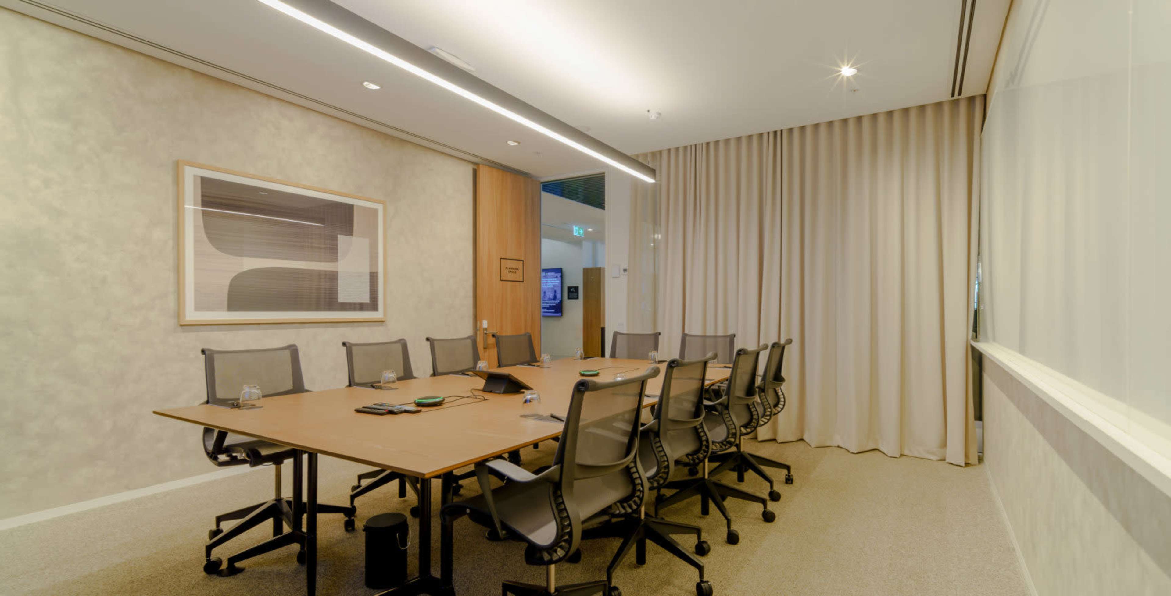 A modern conference room features a large table surrounded by ergonomic chairs, with curtains and a framed artwork along the walls.