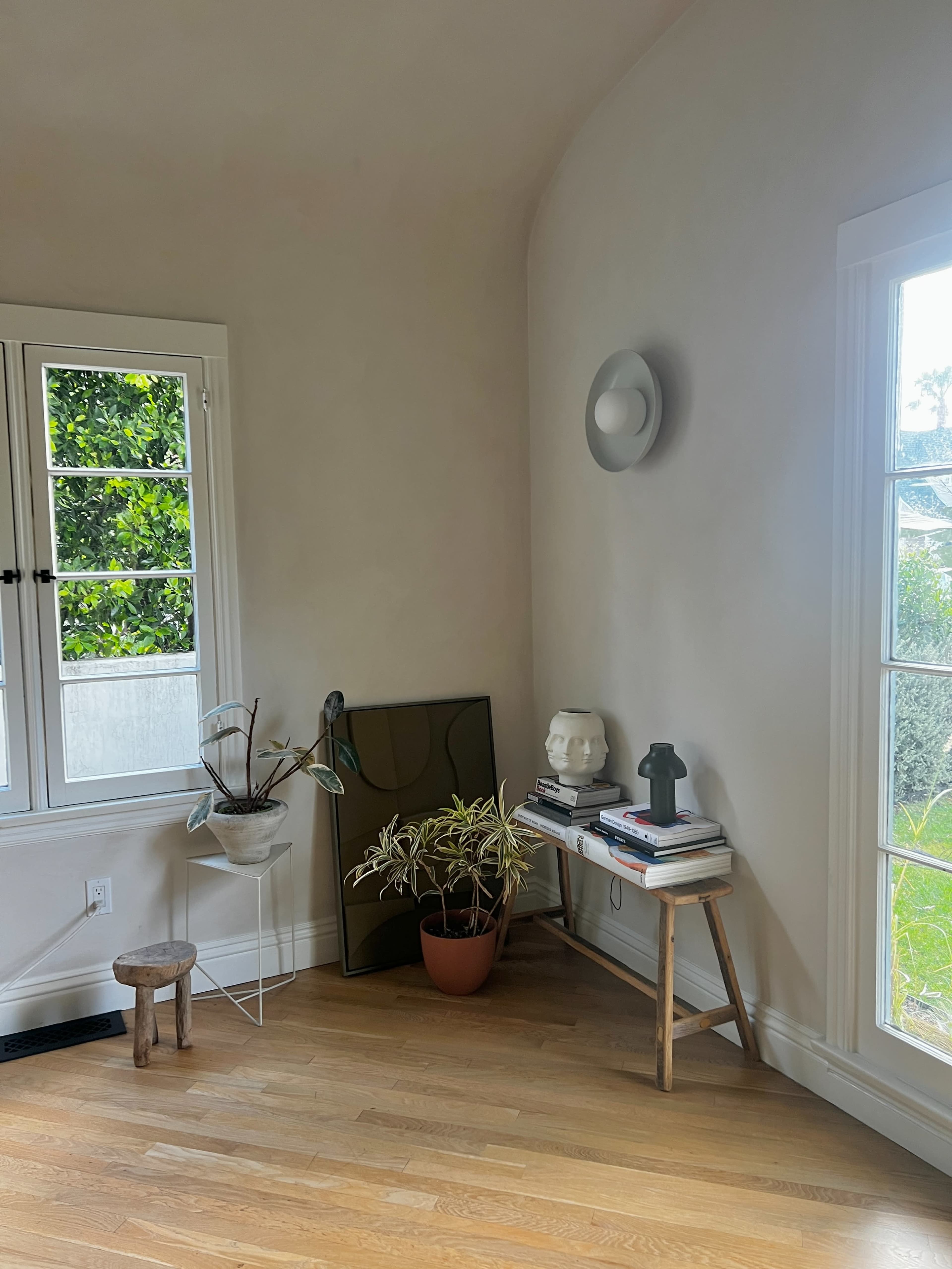 The image shows a cozy corner of a room featuring a plant, a sculpture, and stacked books on a small table beside a large window.