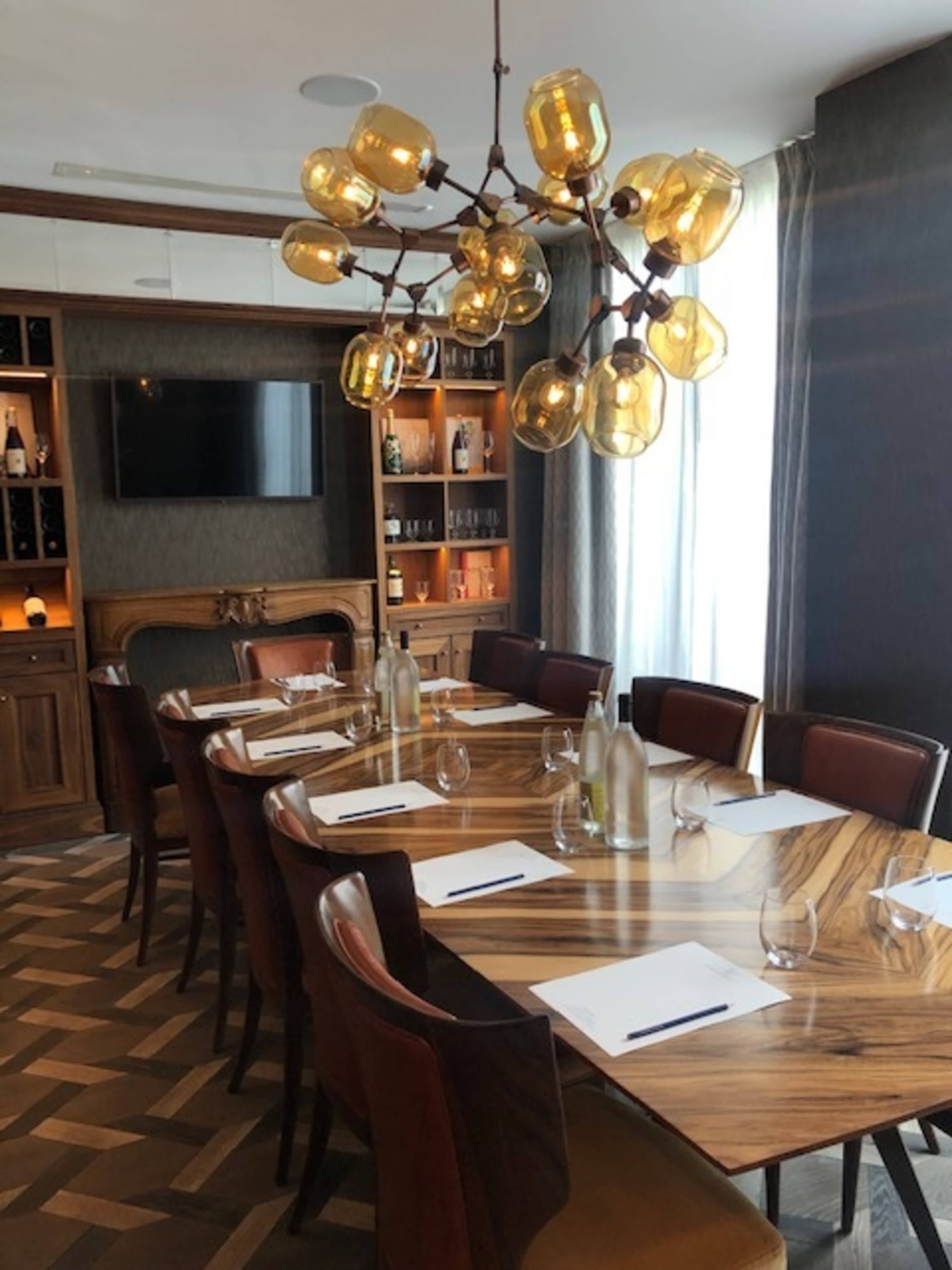 A long wooden dining table is set with glasses and notepads, illuminated by a modern chandelier, in a well-furnished room featuring a wall-mounted TV and shelves with bottles.